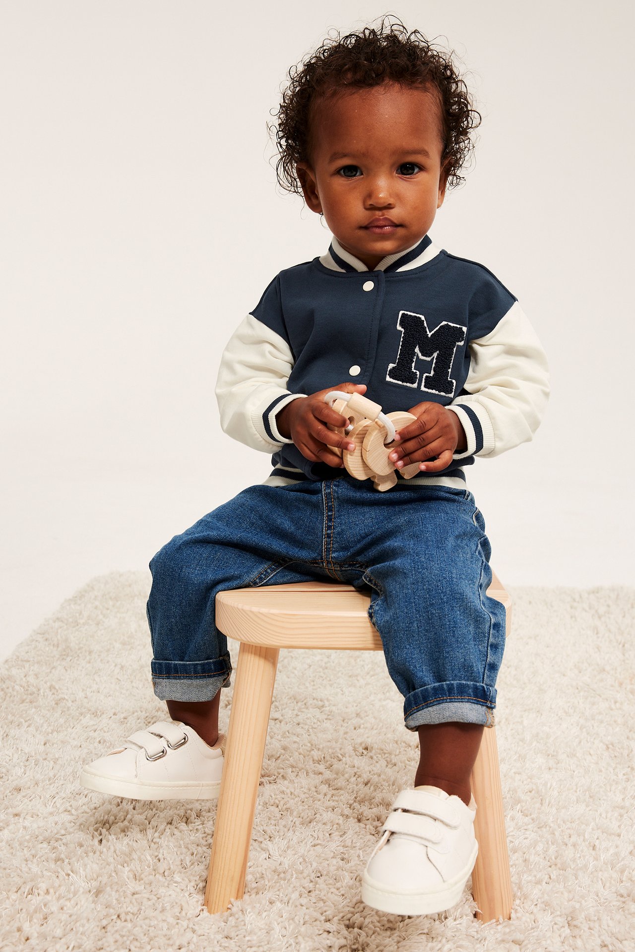 Baby bærer marineblå collegejakke med M-merke, blå jeans, hvite joggesko, holder treleke.