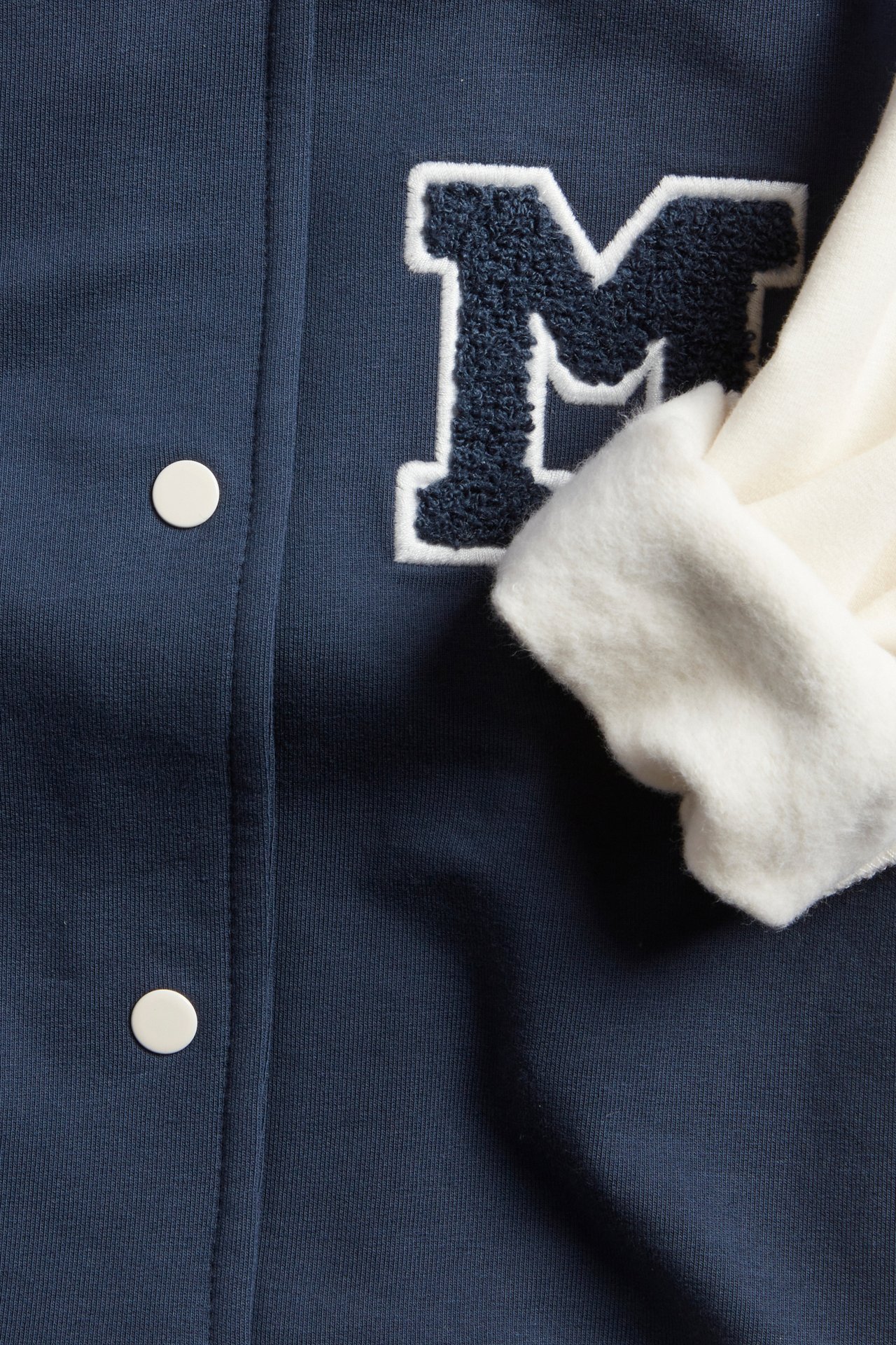 Närbild på en blå baseballjacka med mörkblå M-applikation, vita knappar och en vit ribbad mudd.