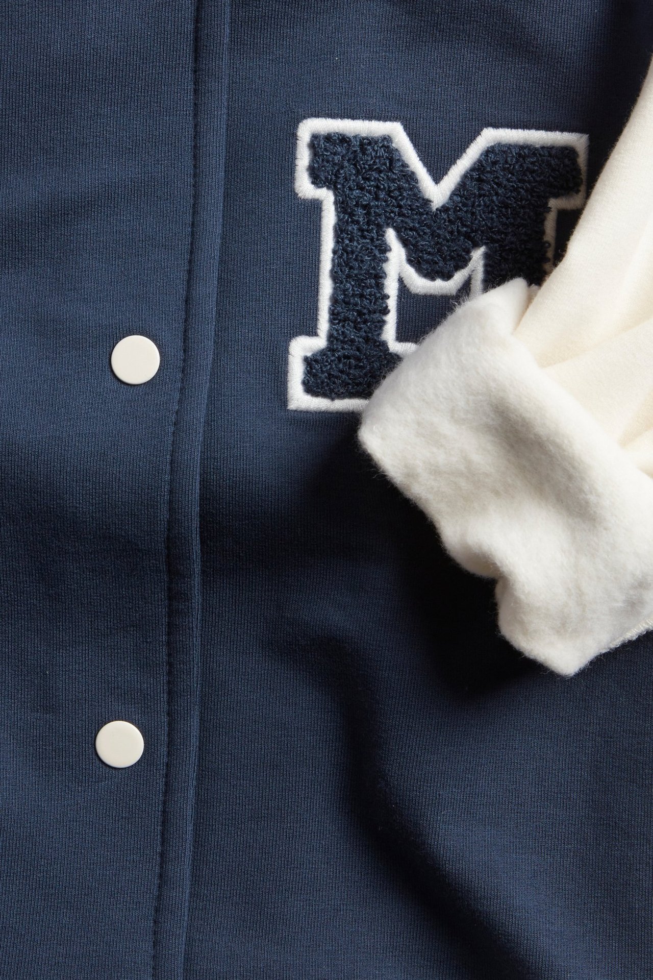 Zbliżenie niebieskiej kurtki bejsbolowej z granatową aplikacją M, białymi guzikami i białym prążkowanym mankietem.