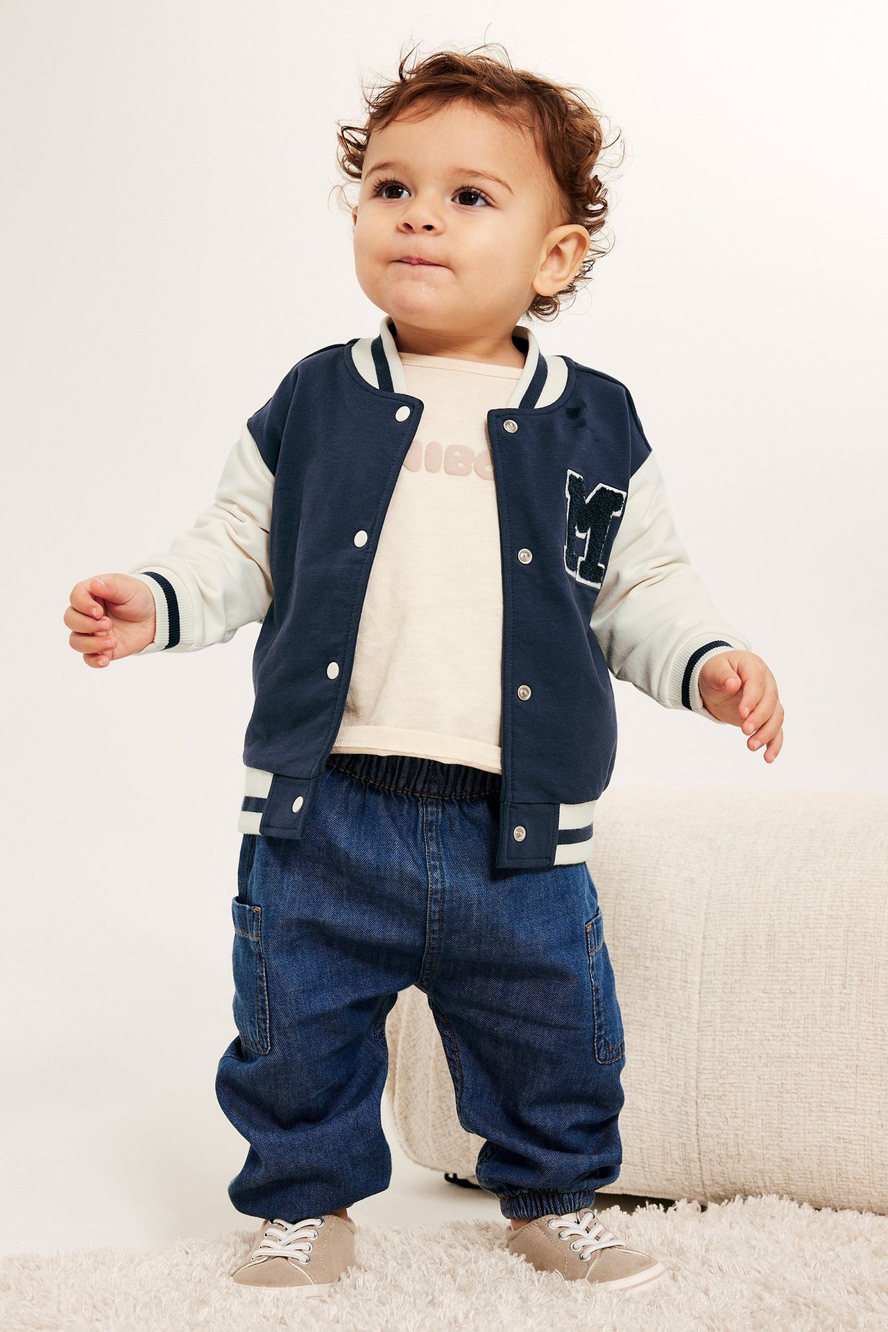 Baby i marinblå och krämfärgad baseballjacka, beige t-shirt, blå jeansjoggers och beige skor.