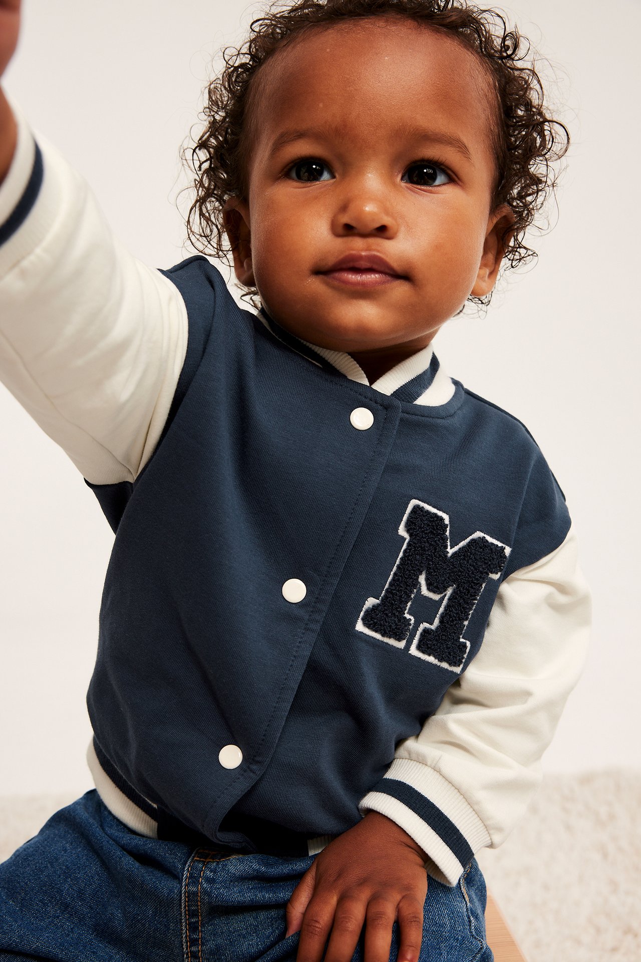 Nærbilde av baby i mørkblå collegejakke med kremfargede ermer og blå denimjeans.