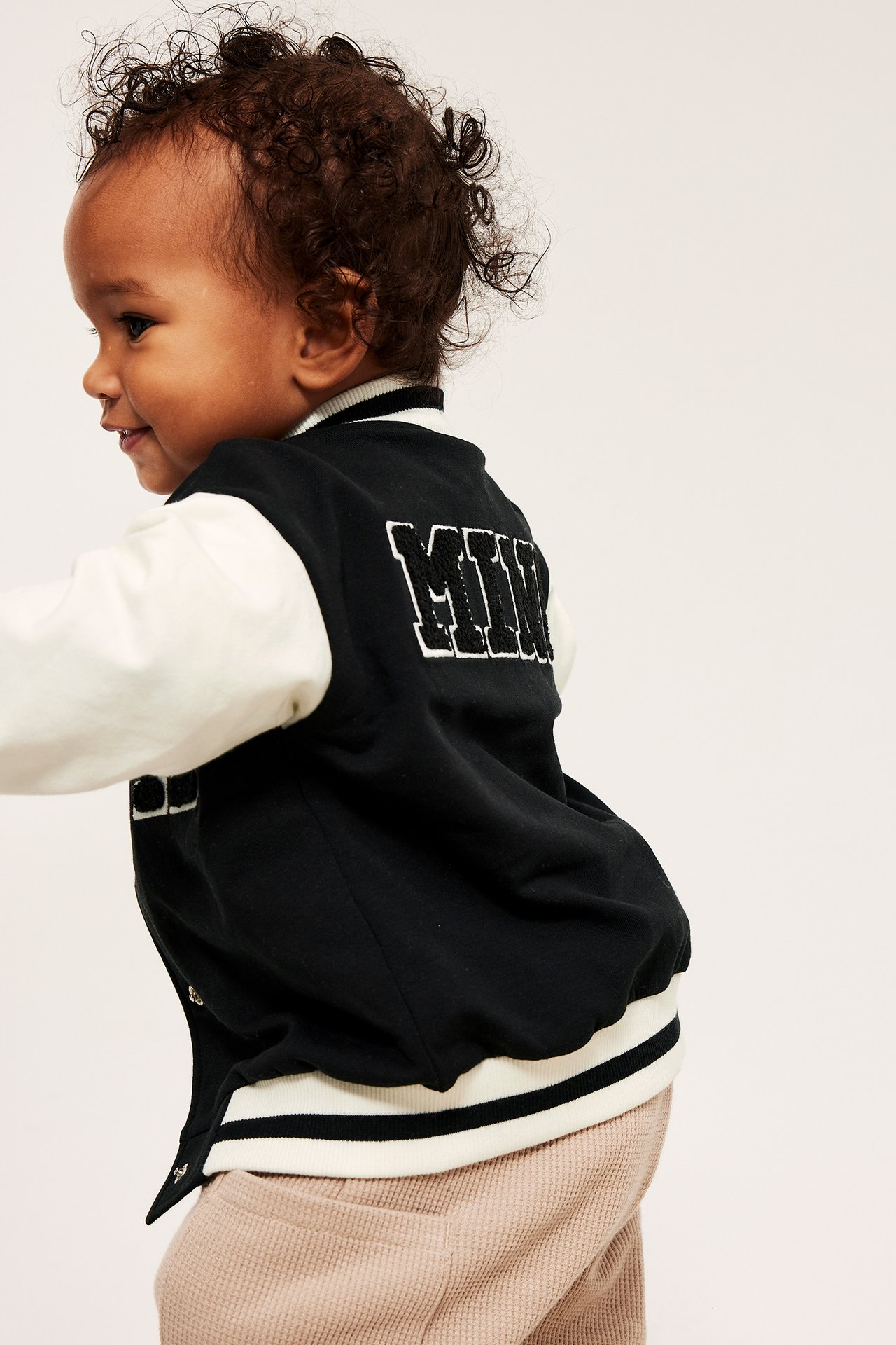 Sned ryggvy av en bebis i svart och vit baseballjacka med brodyr, stylad med beige byxor.