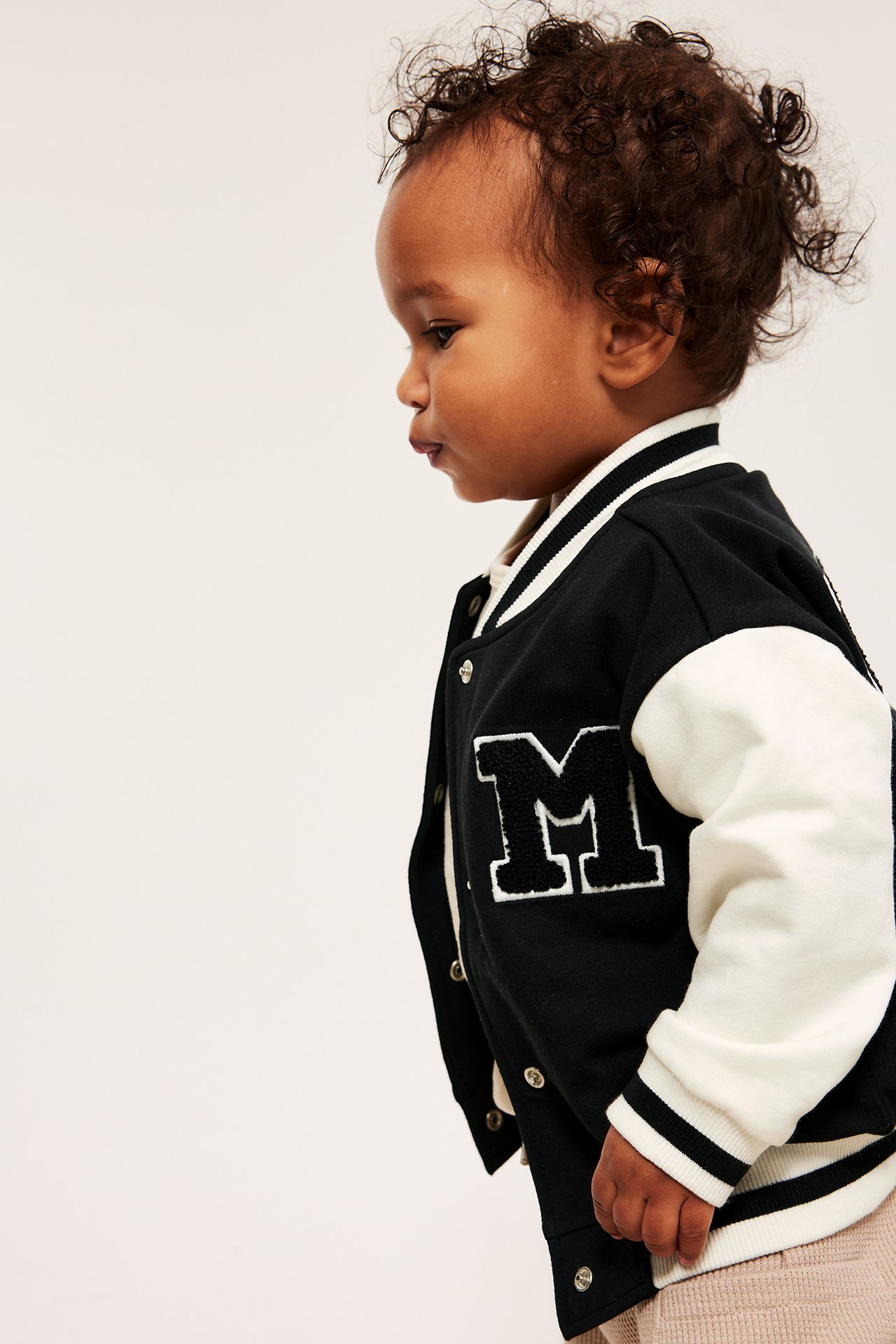 Sivukuva vauvasta yllään mustavalkoinen baseballtakki 'M'-applikaatiolla, ribbiresoreilla ja neppareilla.