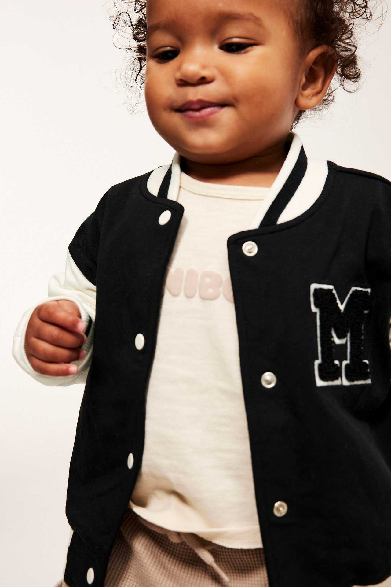 Vauva mustassa baseballtakissa, valkoiset hihat, raidallinen kaulus ja 'M'-applikaatio, napitettu vaalean topin päällä.