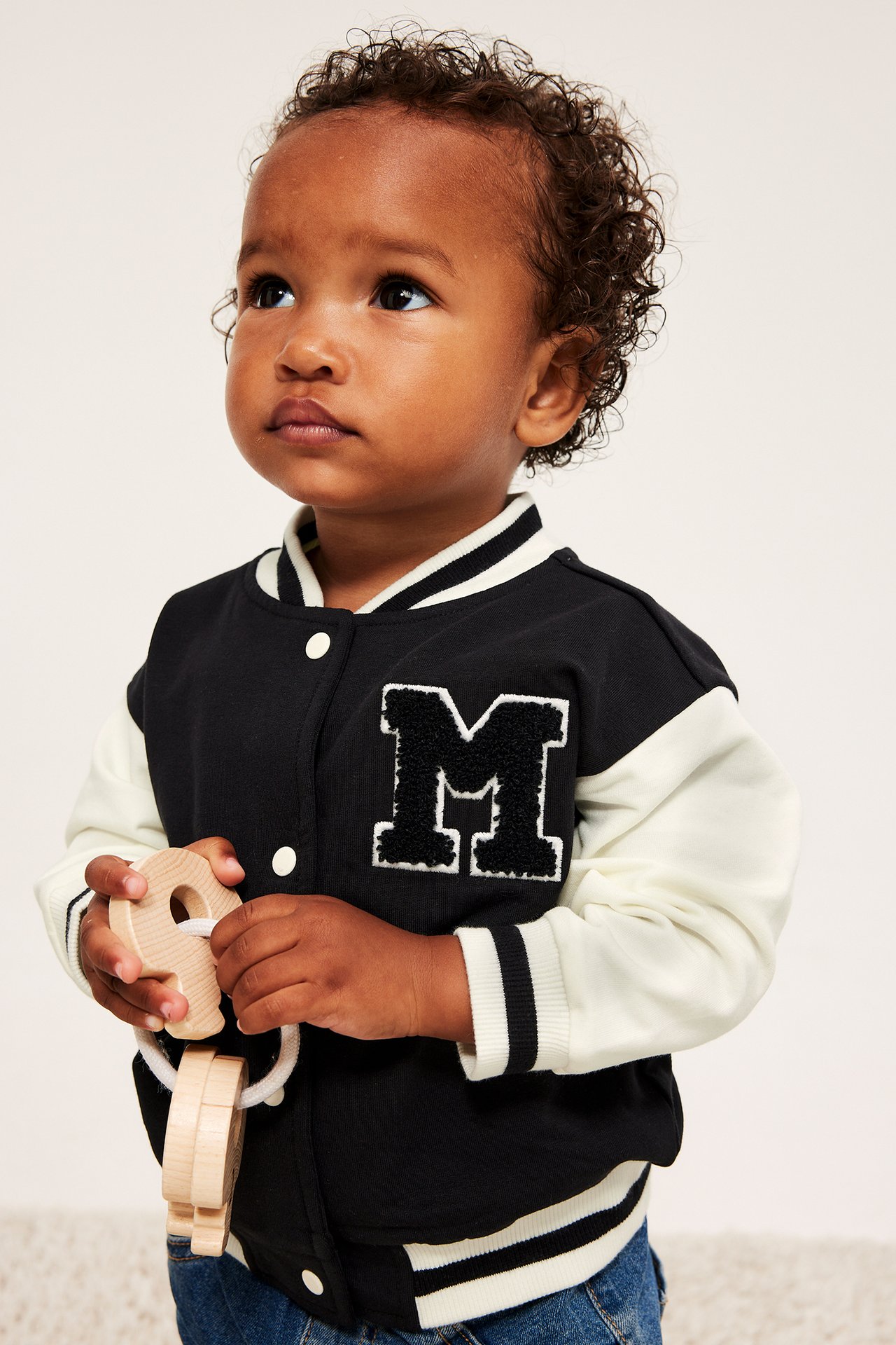 Frontalt bilde av baby i svart og hvit collegejakke med M-emblem, holder treleke.