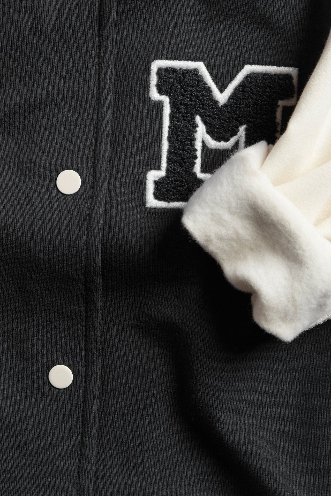 Nærbilde av svart baby baseballjakke med brodert M-merke, kremfarget ermedetalj og hvite trykknapper.