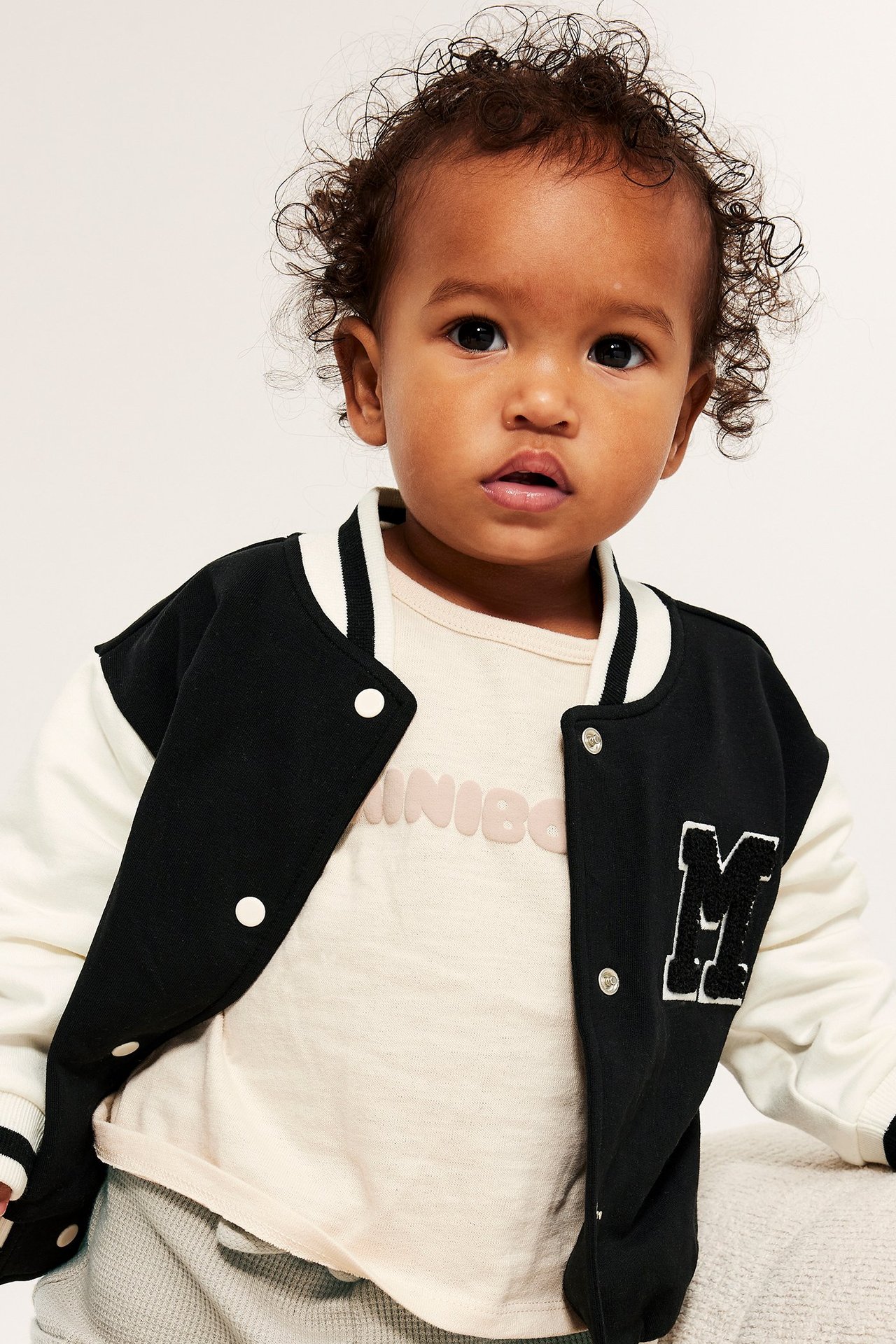 Edestäpäin: Vauva mustassa ja kermanvärisessä baseball-takissa M-applikaatiolla beigen paidan päällä.