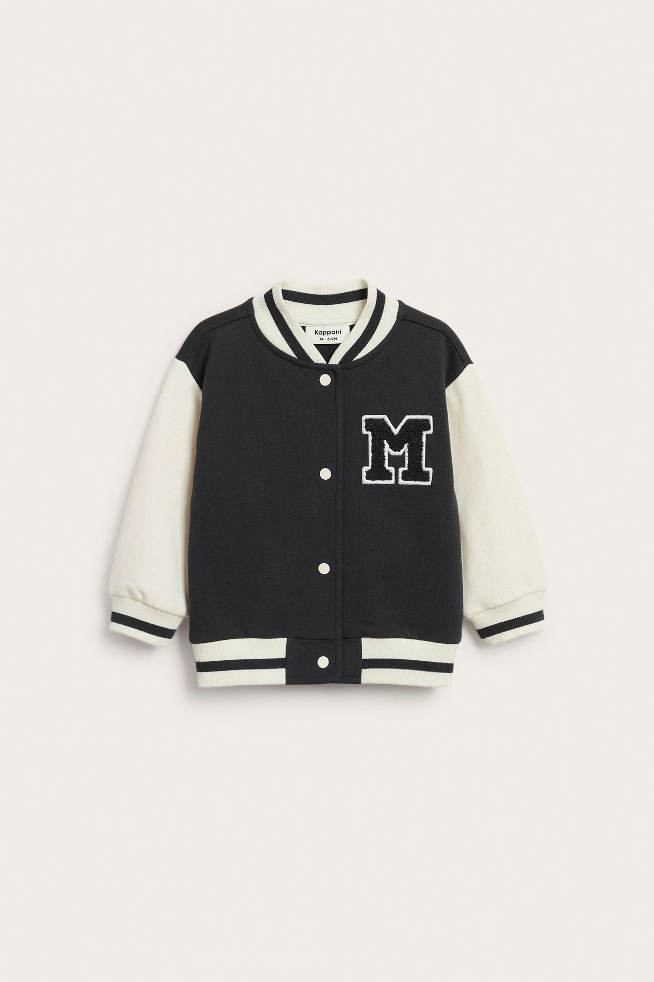 Frontbilde av en babys svart og offwhite baseballjakke med hvit 'M'-applikasjon og trykknapper.