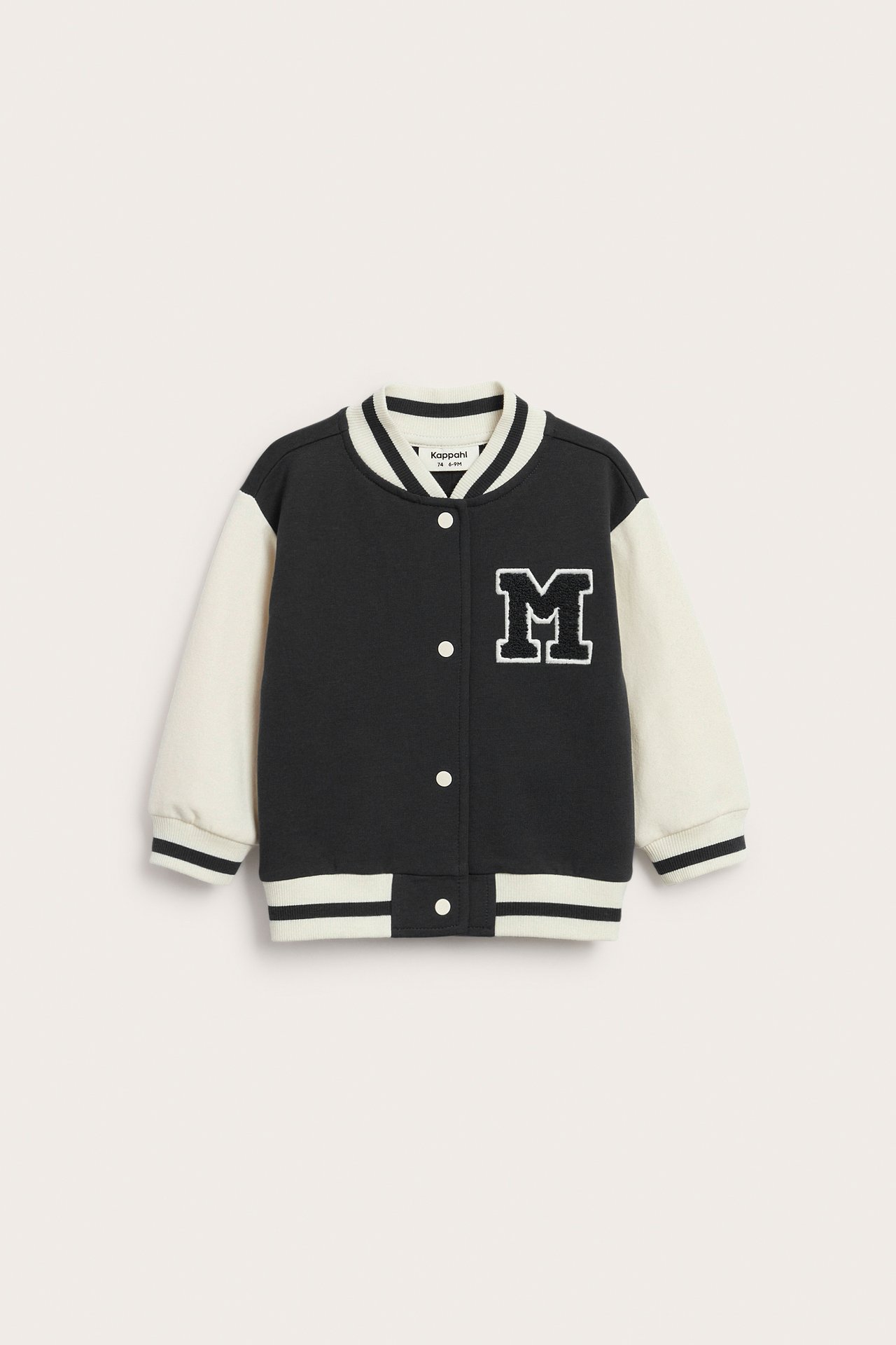 Front av barn varsityjakke i svart og off-white med M-patch og trykknapper.