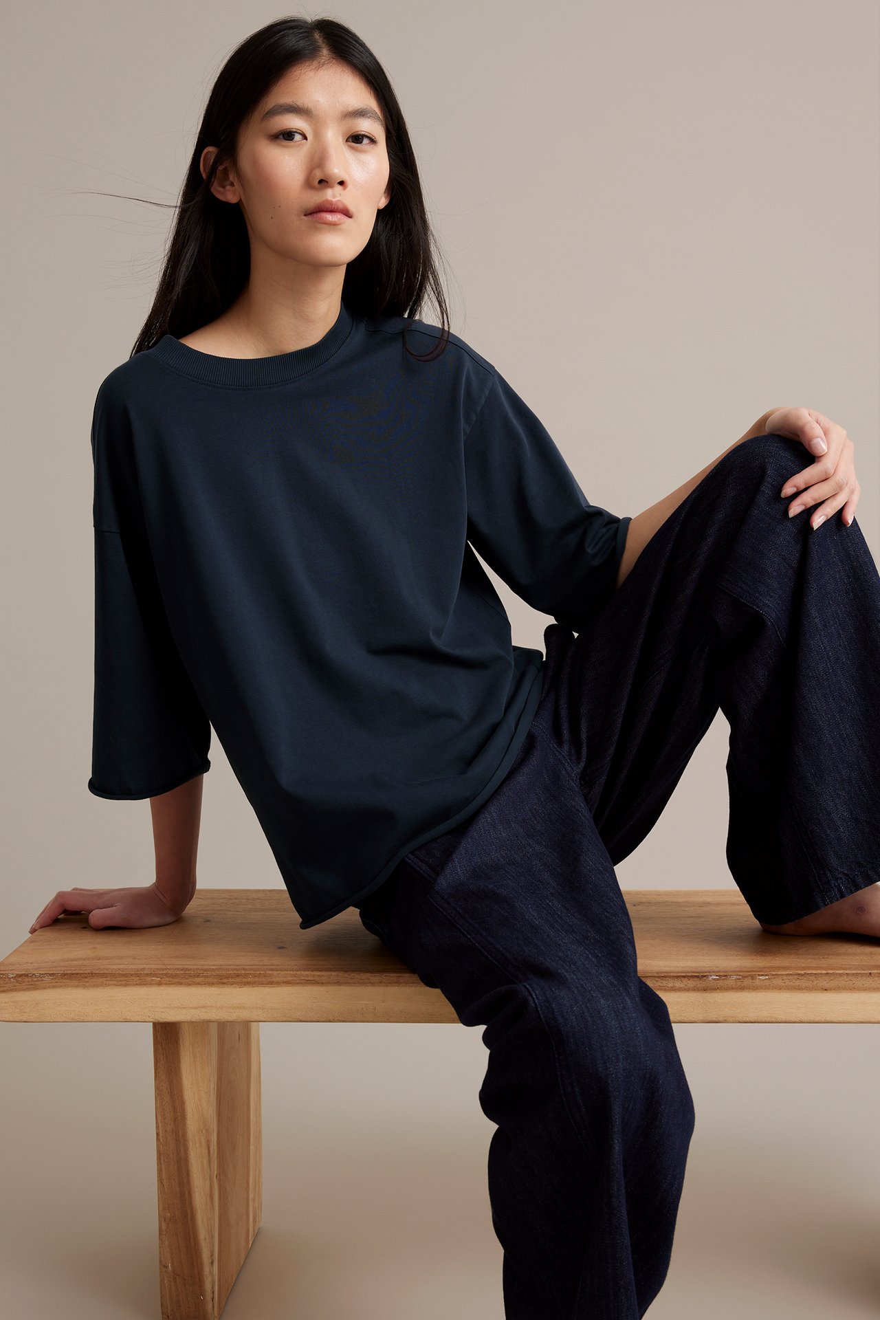 Kvinna i mörkblå T-shirt med råa fållar, loose fit, framför, sittande på träbänk.