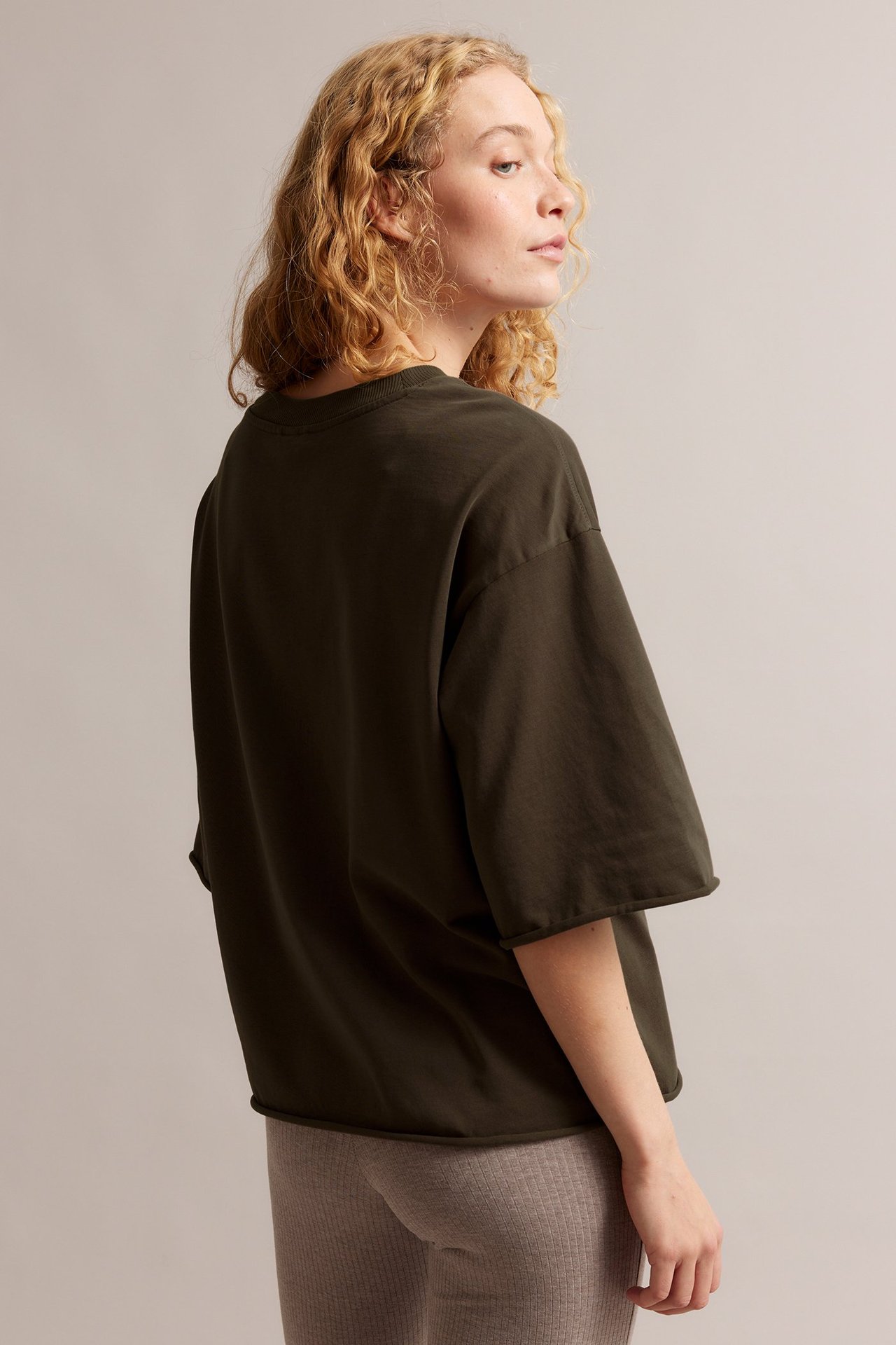 Bakifrån, en kvinna i en lös mörkbrun bomulls-t-shirt med råa fållar över ljusa ribbade leggings.
