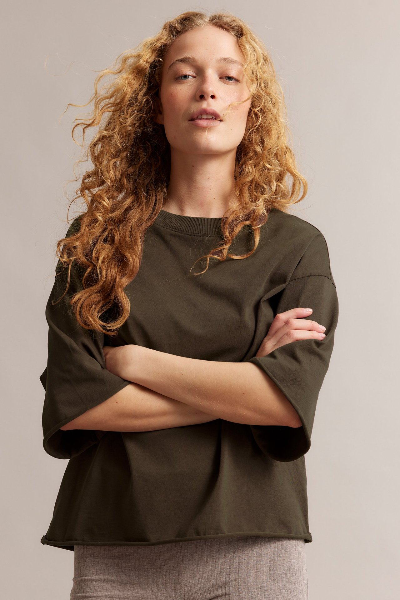 Framifrån, kvinna bär en lös brun T-shirt i bomull med råa fållar över ljusa ribbstickade byxor.