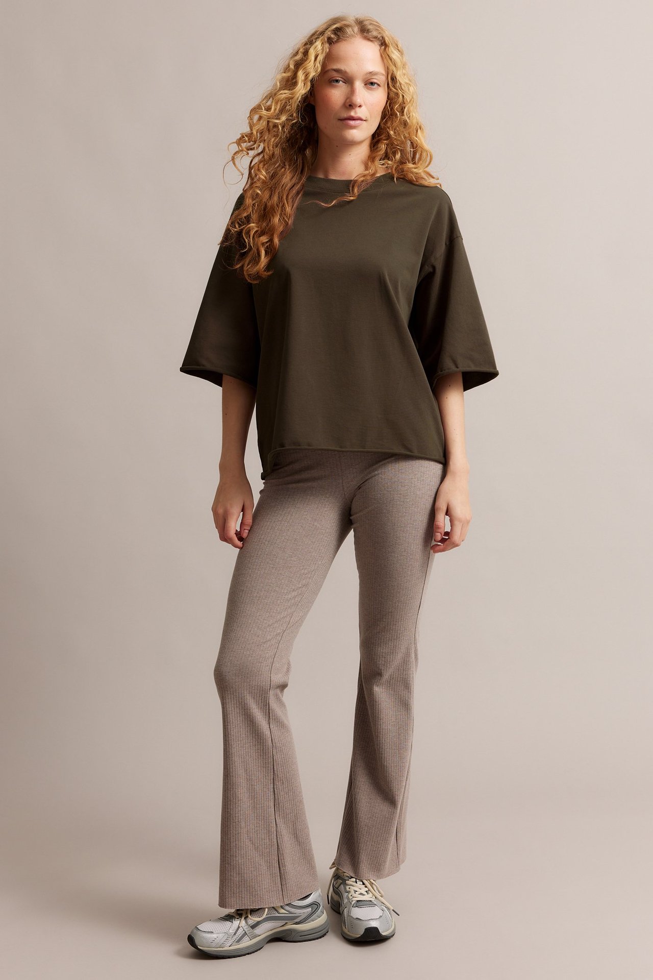Edestä: Nainen pukeutuneena löysään tummanruskeaan puuvilla T-paitaan, vaaleanruskeisiin kuviollisiin leveneviin housuihin ja harmaisiin lenkkareihin.
