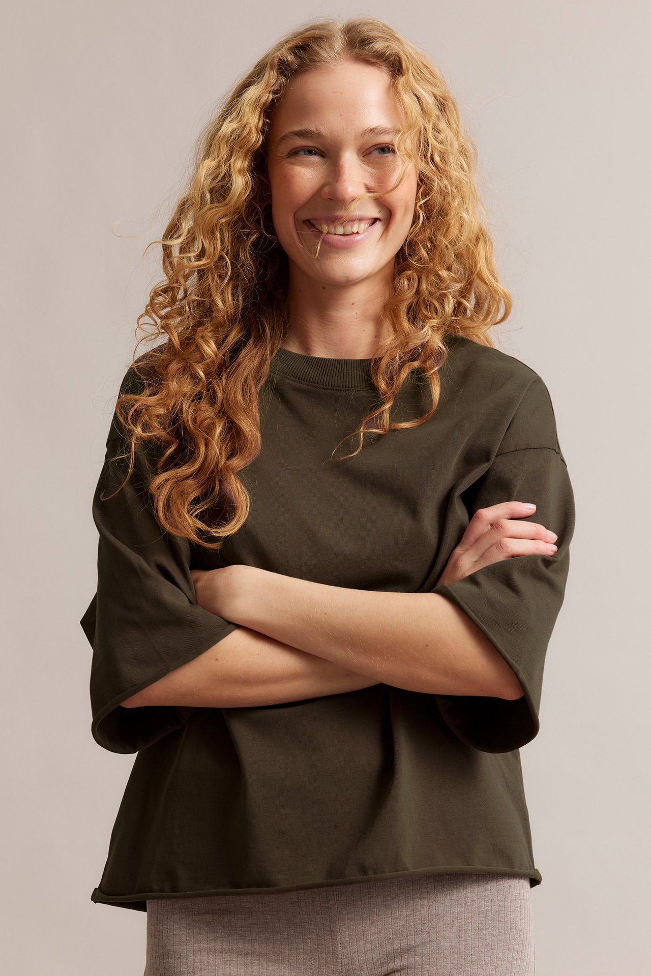 Uśmiechnięta kobieta, z przodu, w ciemnozielonej luźnej koszulce z surowymi brzegami i jasnobrązowych legginsach.
