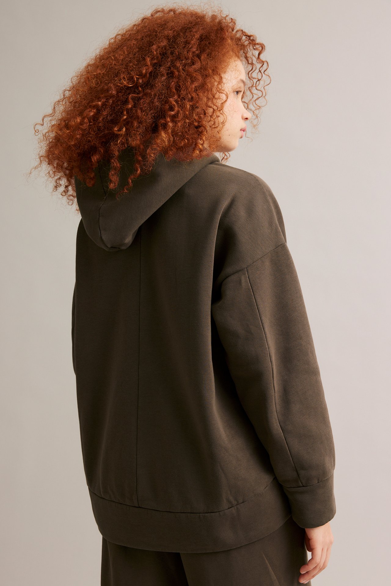 Naisen taka-sivukuva: tummanruskea yksivärinen oversized huppari huppu ylhäällä ja yhteensopivat alaosat.