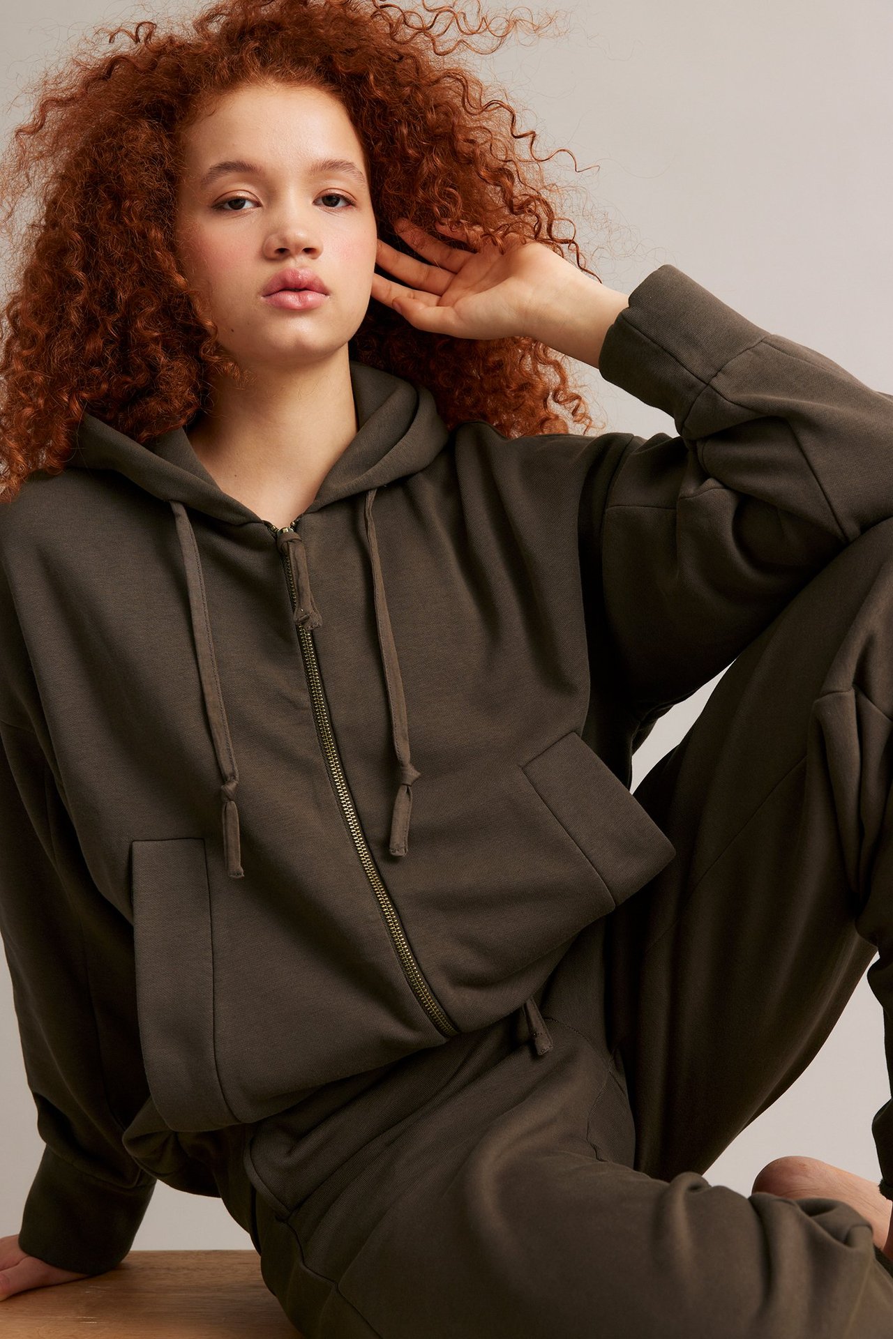 Frontbilde av kvinne i mørk brun hettegenser med glidelås og hette, pluss matchende joggebukse.