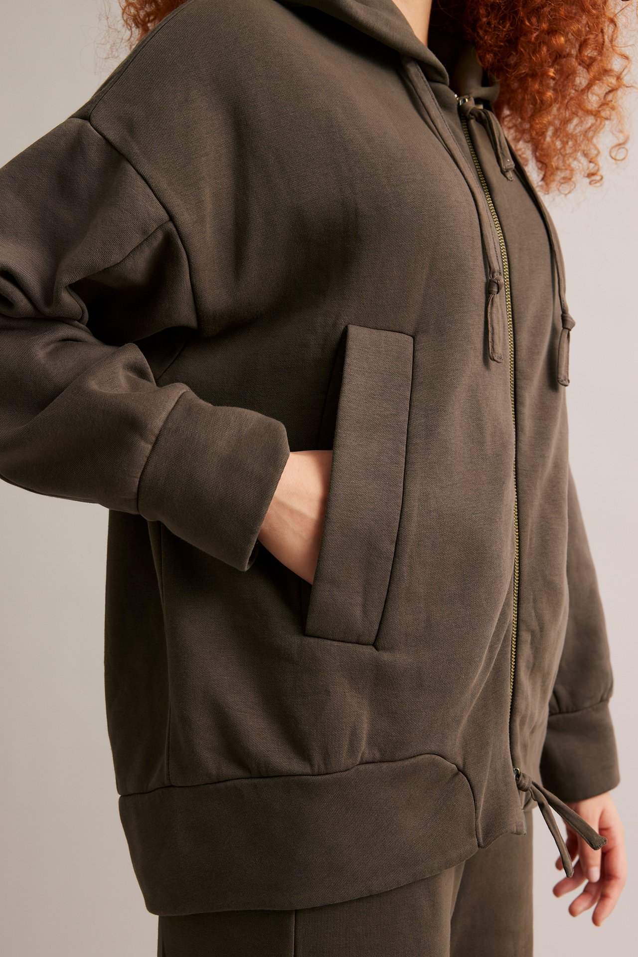Nærbilde sideprofil av en kvinne i en mørkebrun zip-up hettegenser med frontlommer og matchende joggebukse.