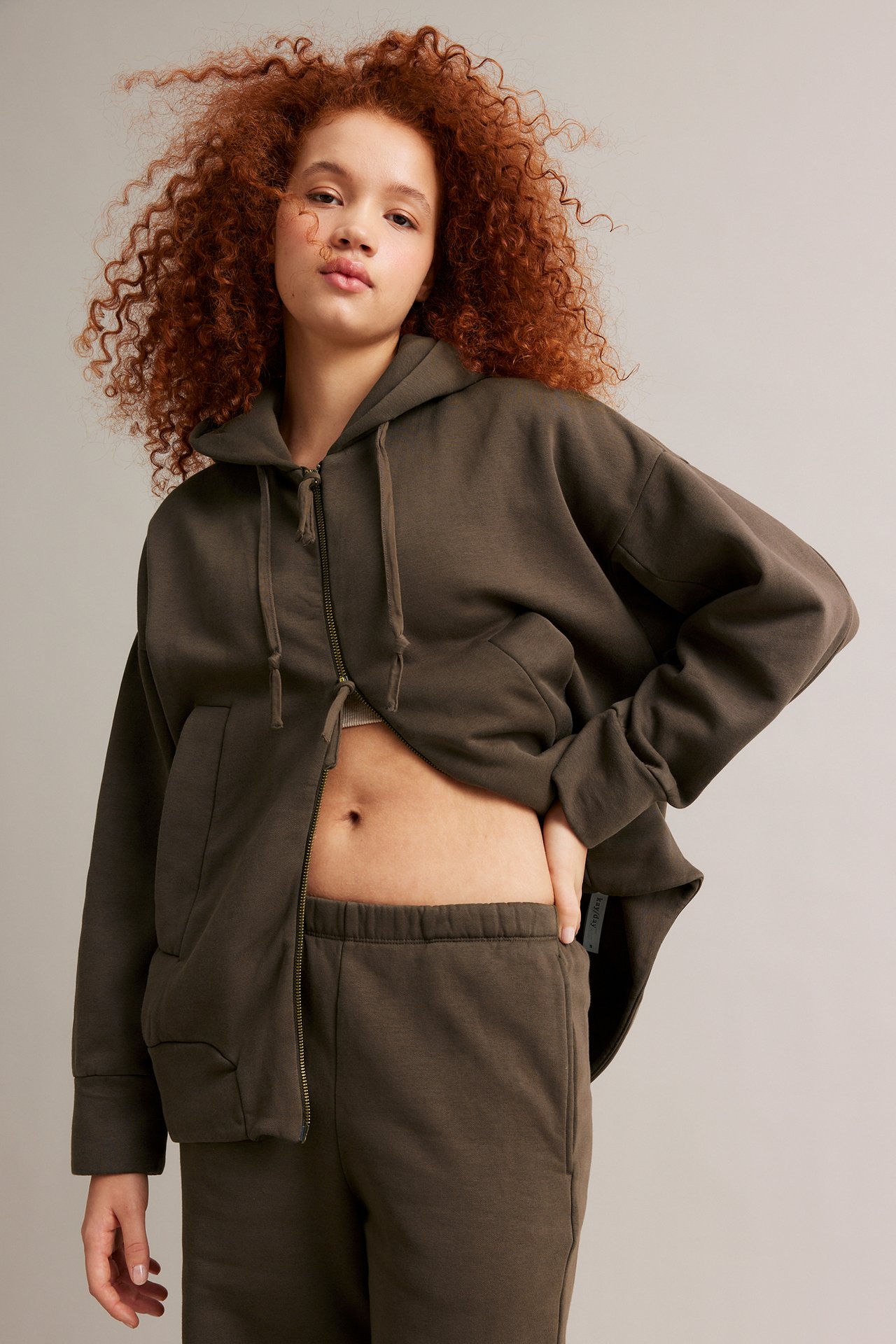 Forfra, en kvinne i åpen brun hettegenser over lys topp og matchende joggebukse.