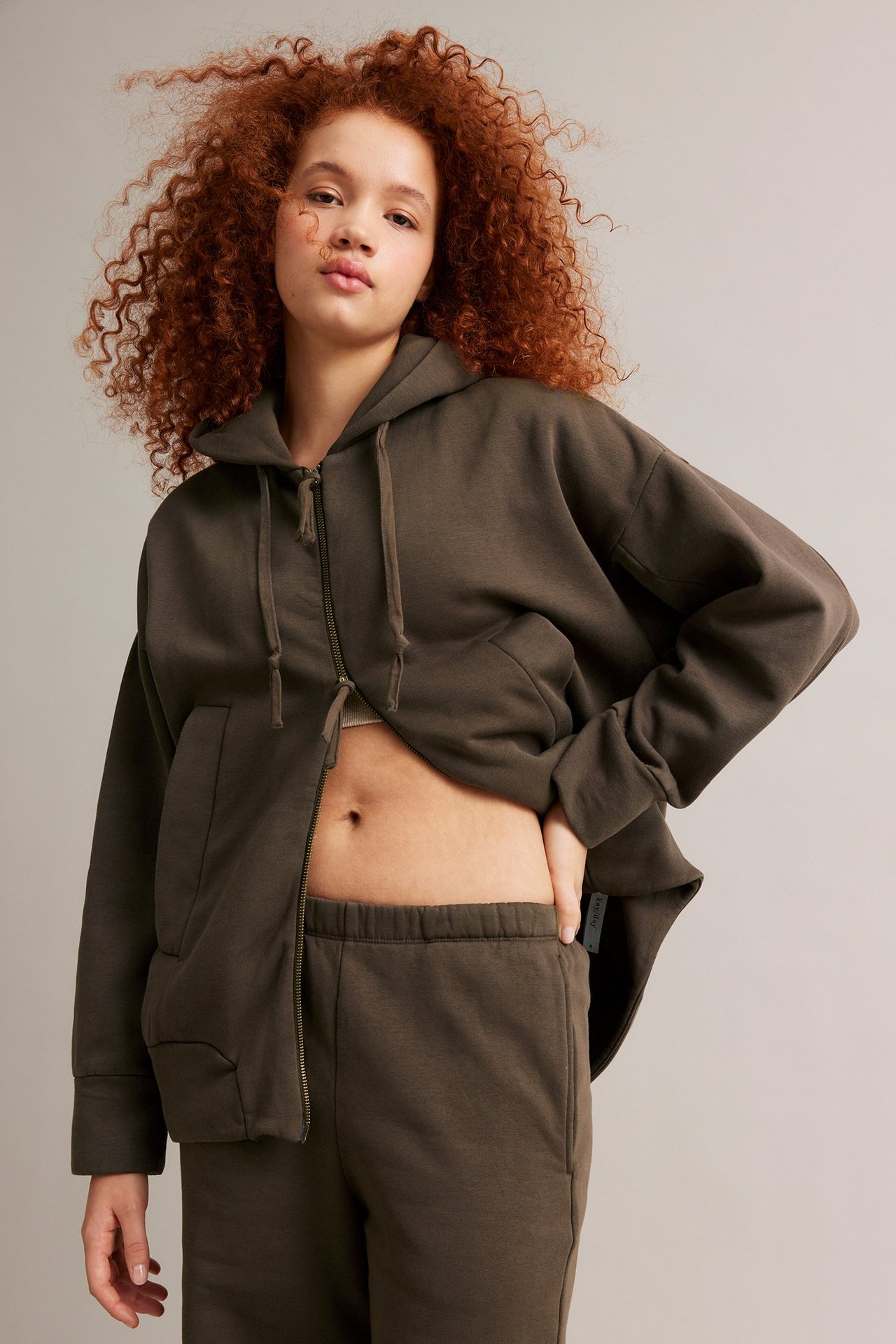 Forfra, en kvinne i åpen brun hettegenser over lys topp og matchende joggebukse.