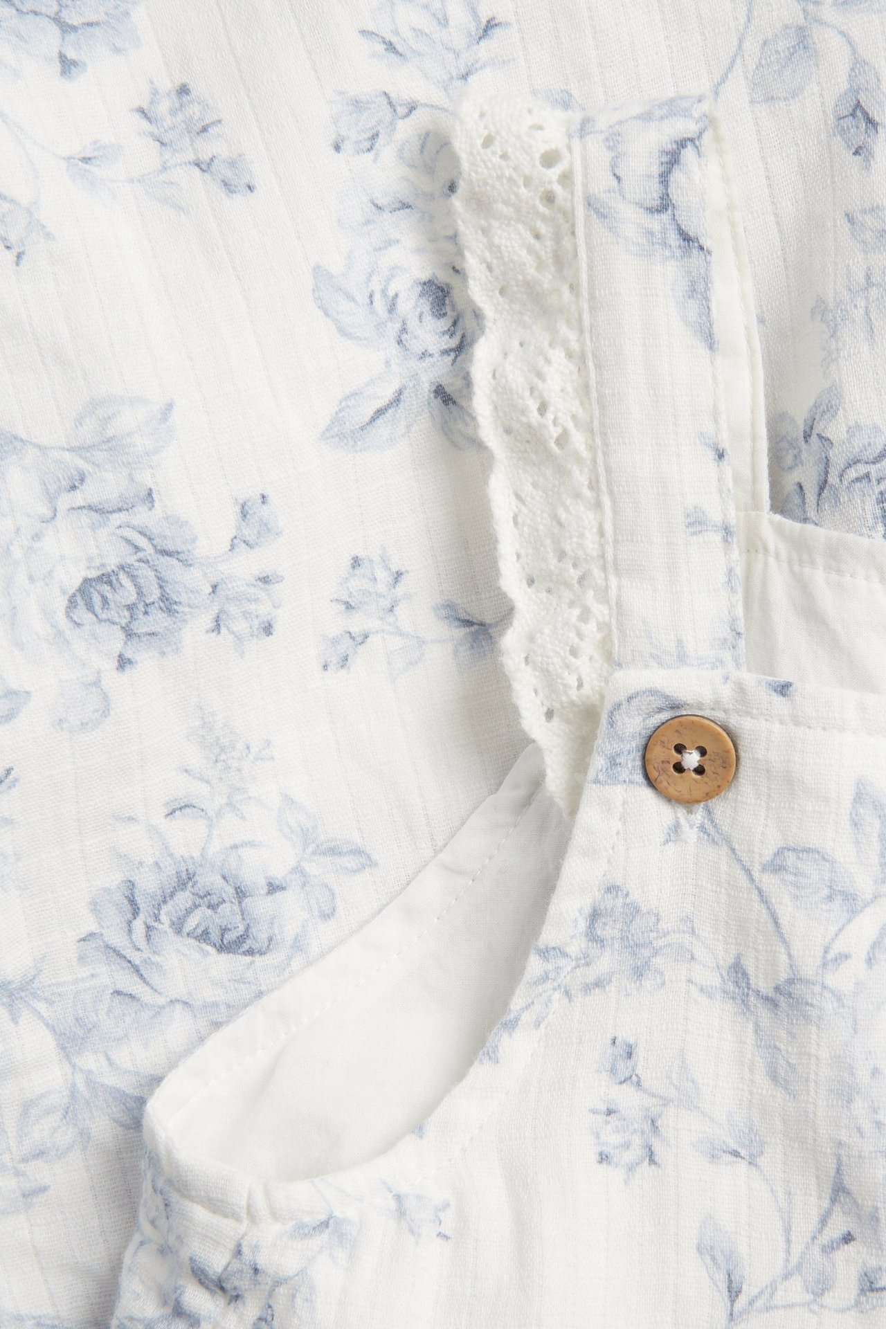 Zbliżenie na off-white bawełniany kombinezon w niebieskie kwiaty, z koronkową falbaną i drewnianym guzikiem.