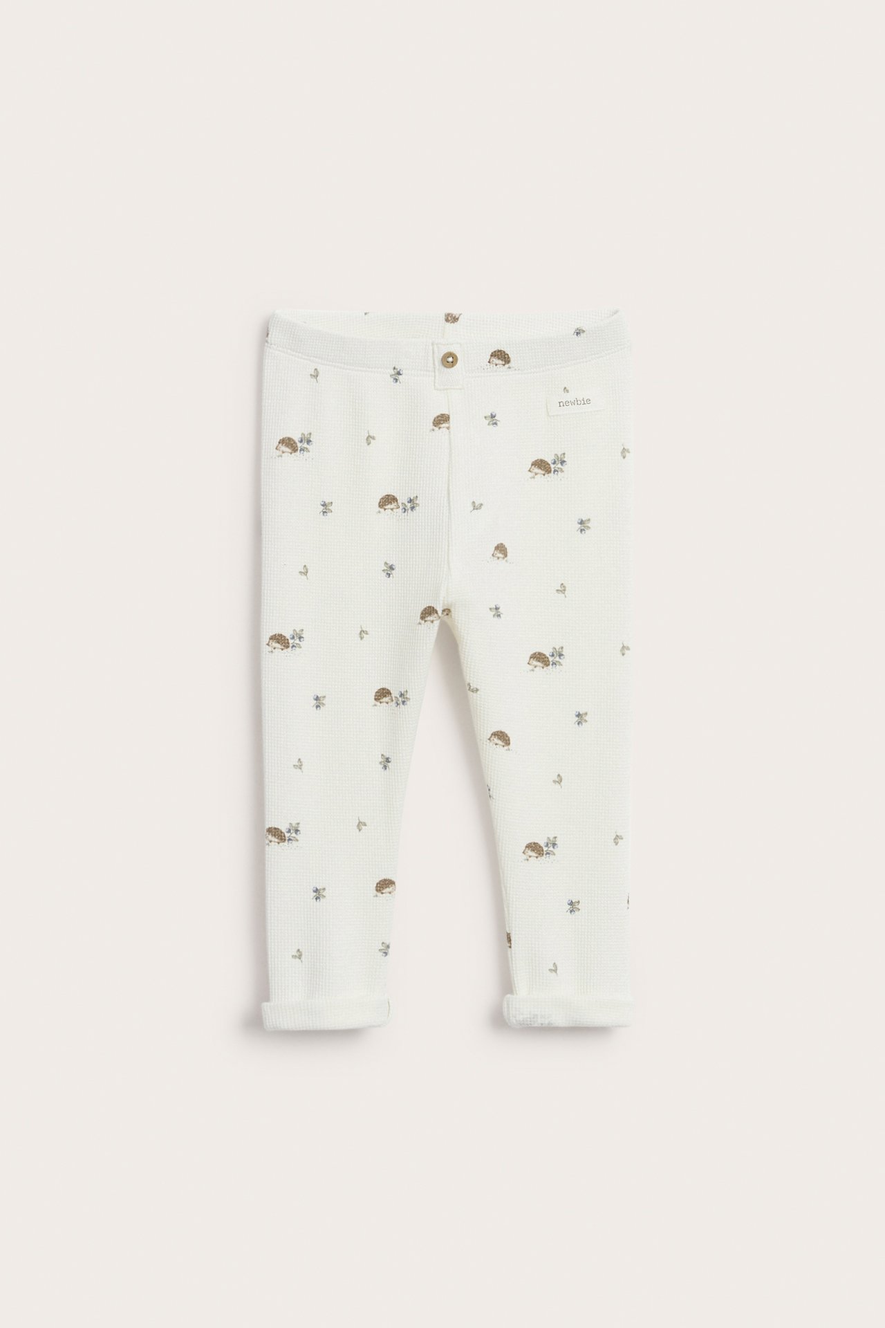 Framifrån: Offwhite våffelstickade baby leggings med igelkottsmönster, dekorativ knapp fram och upprullade benslut.