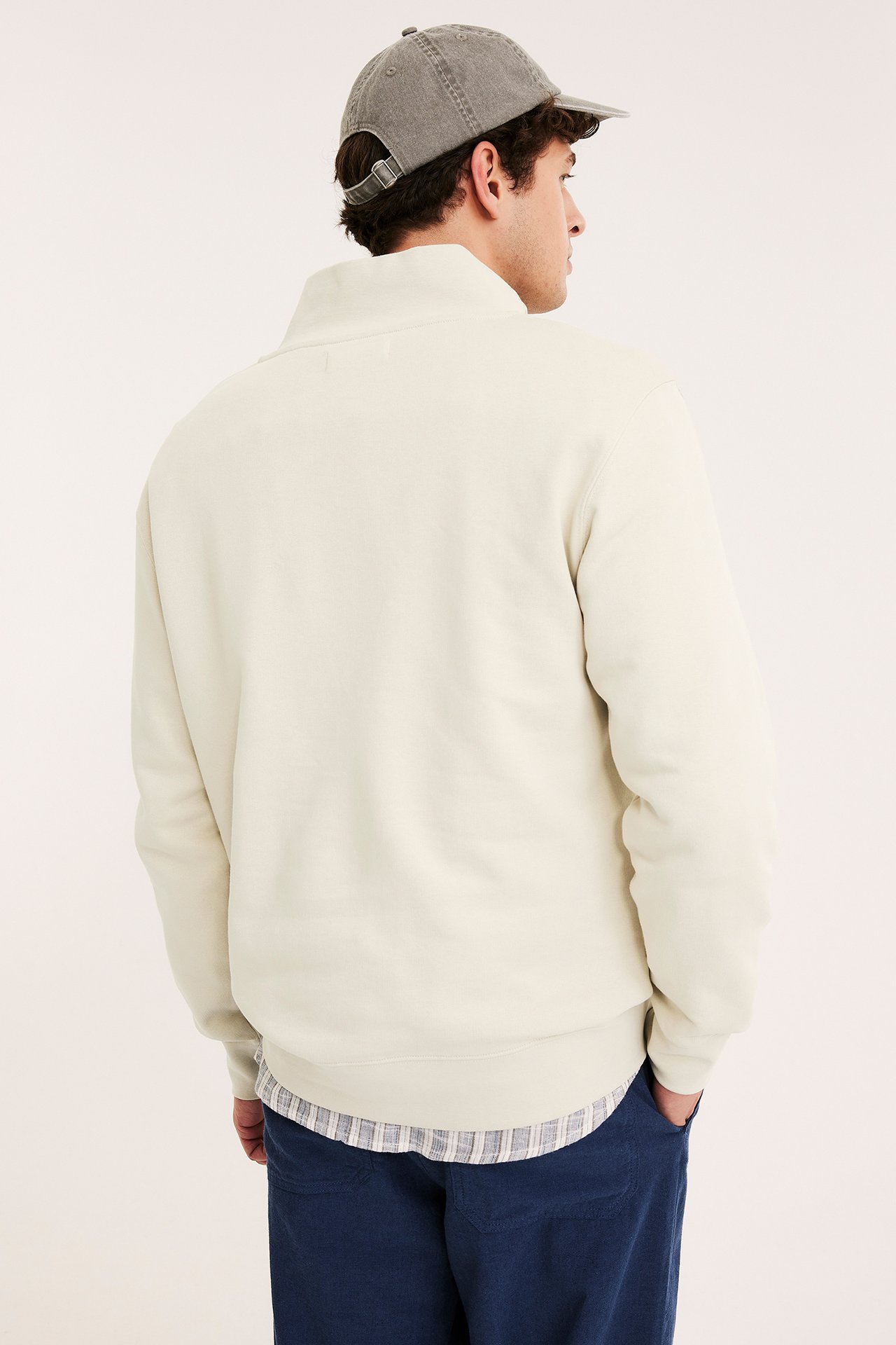 Widok z tyłu: Mężczyzna w jasnobeżowej bluzie z zamkiem, kraciastej koszuli, szarej czapce i niebieskich spodniach.