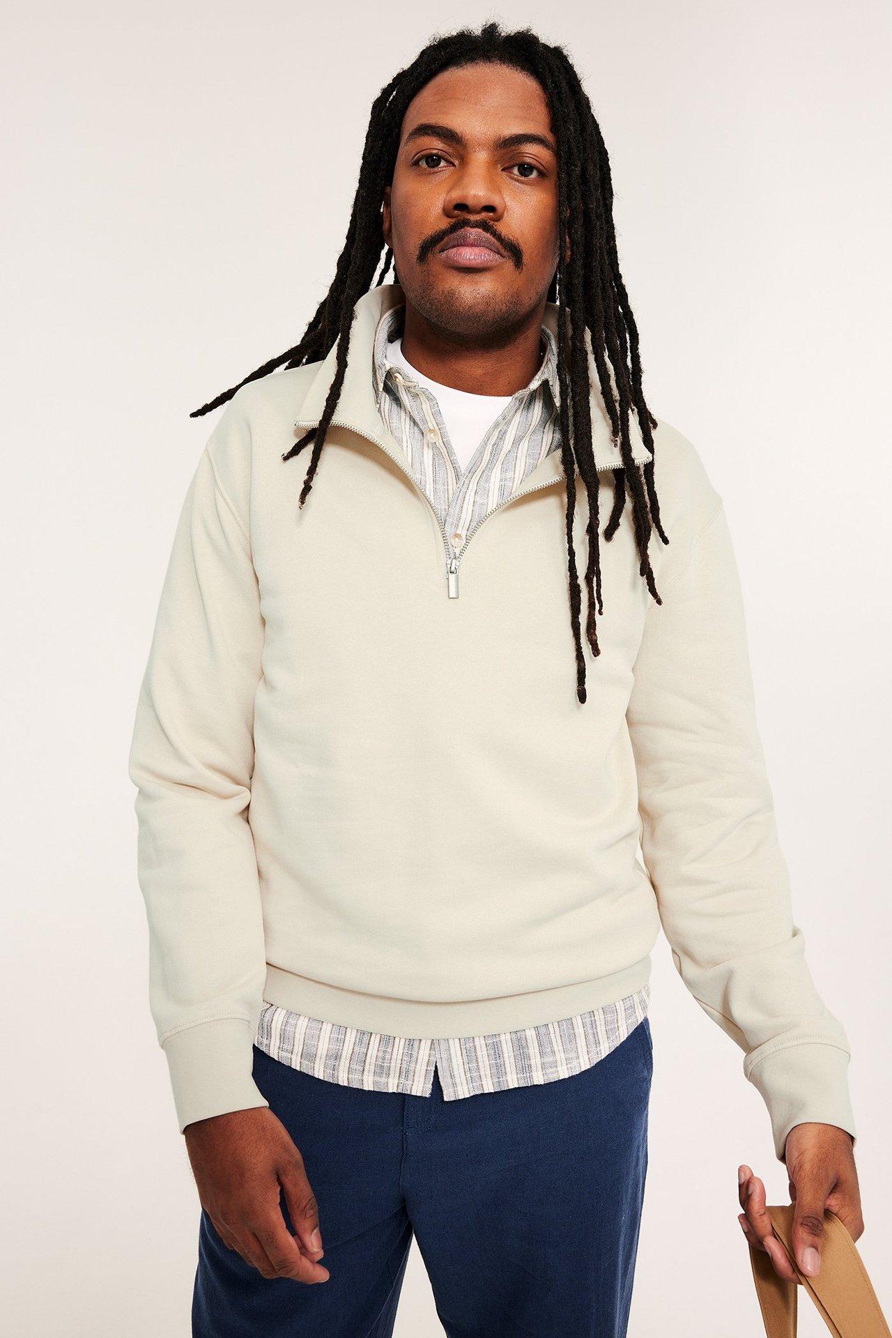 Mann i lys beige half-zip sweatshirt over stripete skjorte og mørkeblå bukser, holder en stropp.