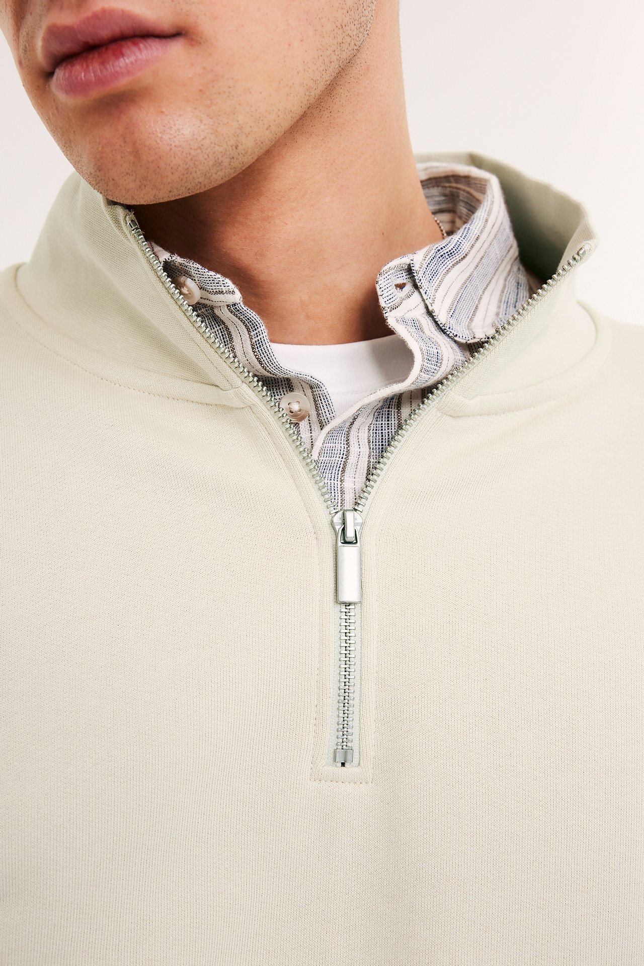 Zbliżenie mężczyzny w jasnobeżowej bluzie z zamkiem do połowy i wysokim kołnierzem, na koszuli w paski.