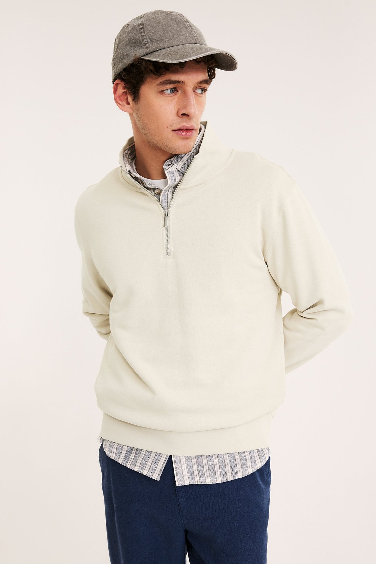 Frontvy av en man i ljusbeige halvziptröja över en rutig skjorta, mörkblå byxor och en grå keps.