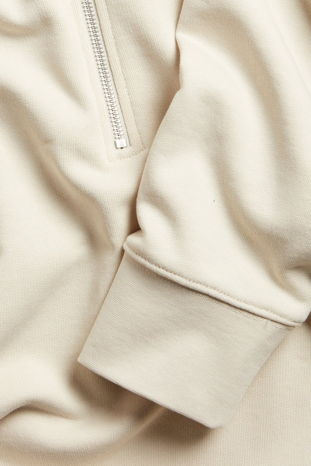 Nærbilde av en lys beige bomullssweatshirt med sølvfarget halv glidelås og ribbet mansjett.