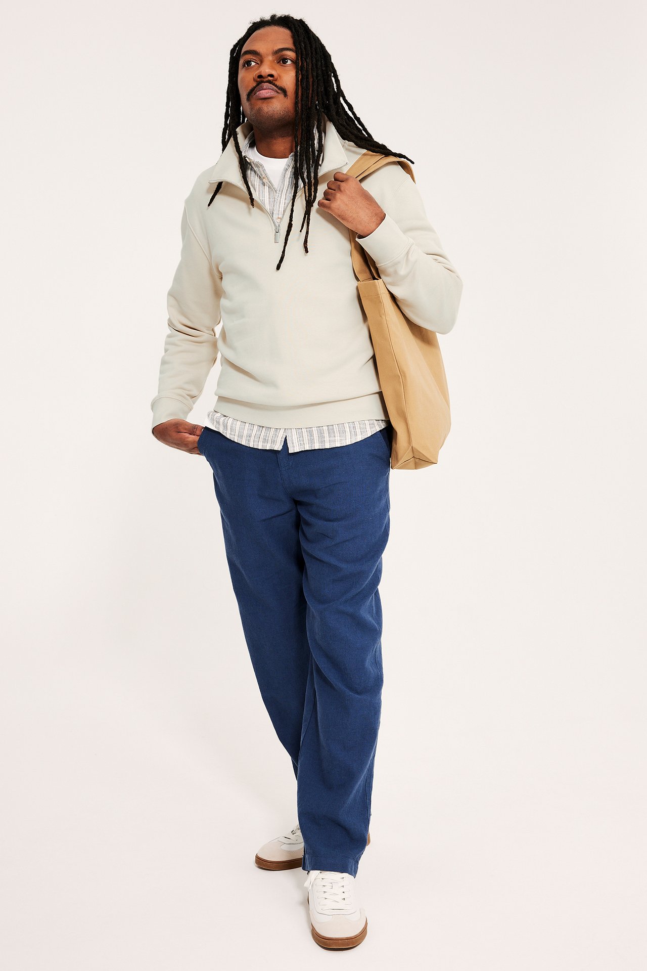 Mann har på seg en lys beige half-zip sweatshirt over stripet skjorte, blå bukser, hvite joggesko, med en beige tote bag.