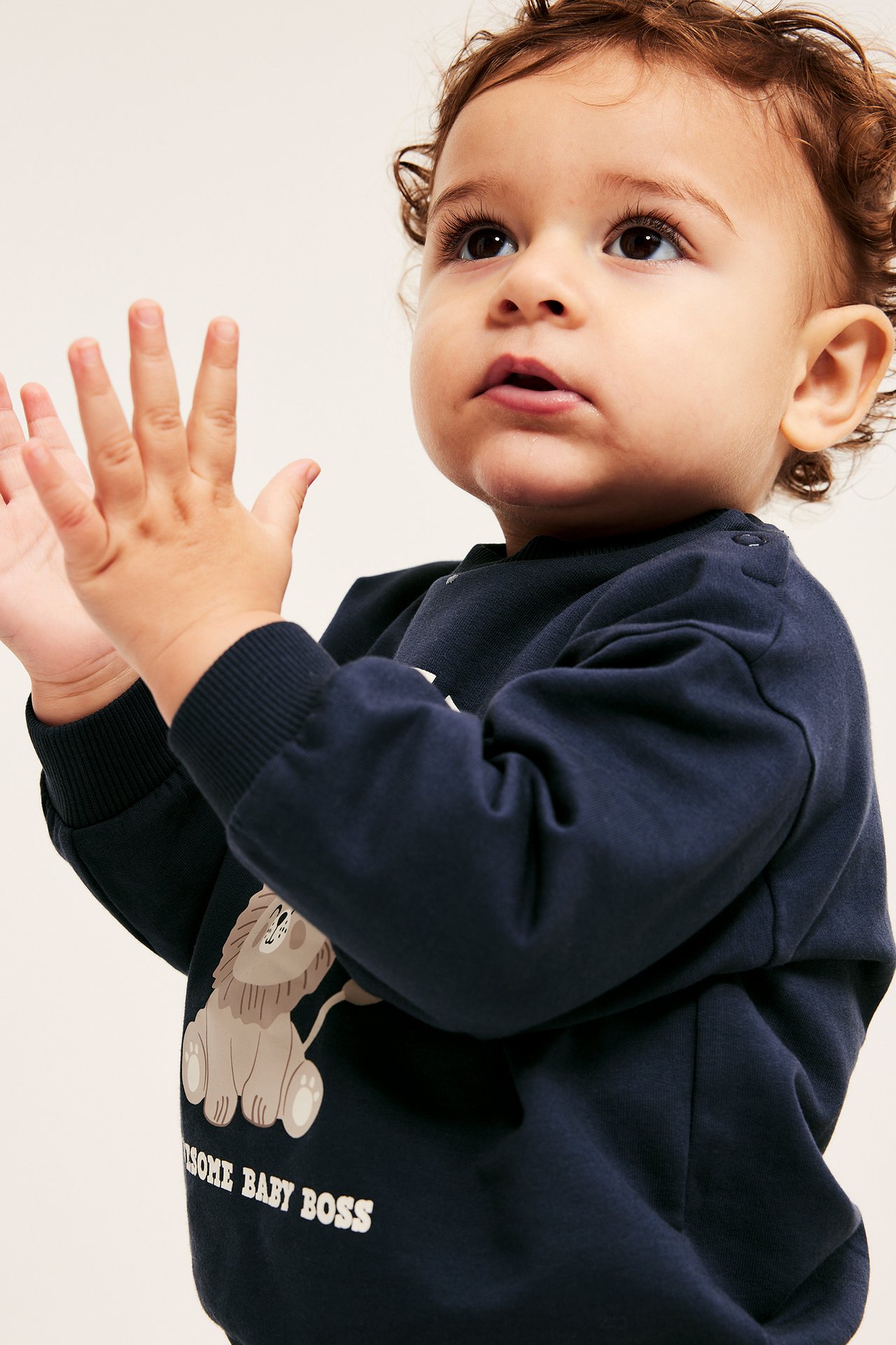 Nærbilde av baby i mørkeblå bomullssweatshirt med løvetrykk, trykknapper på skulderen og ribbestrikkede mansjetter.