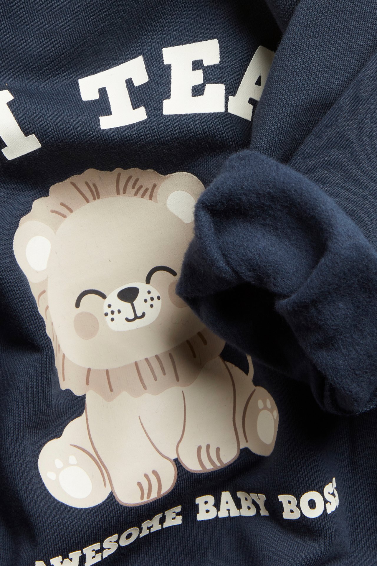 Nærbilde av en mørkeblå babysweater med smilende løvetrykk og ribbestrikkede mansjetter.