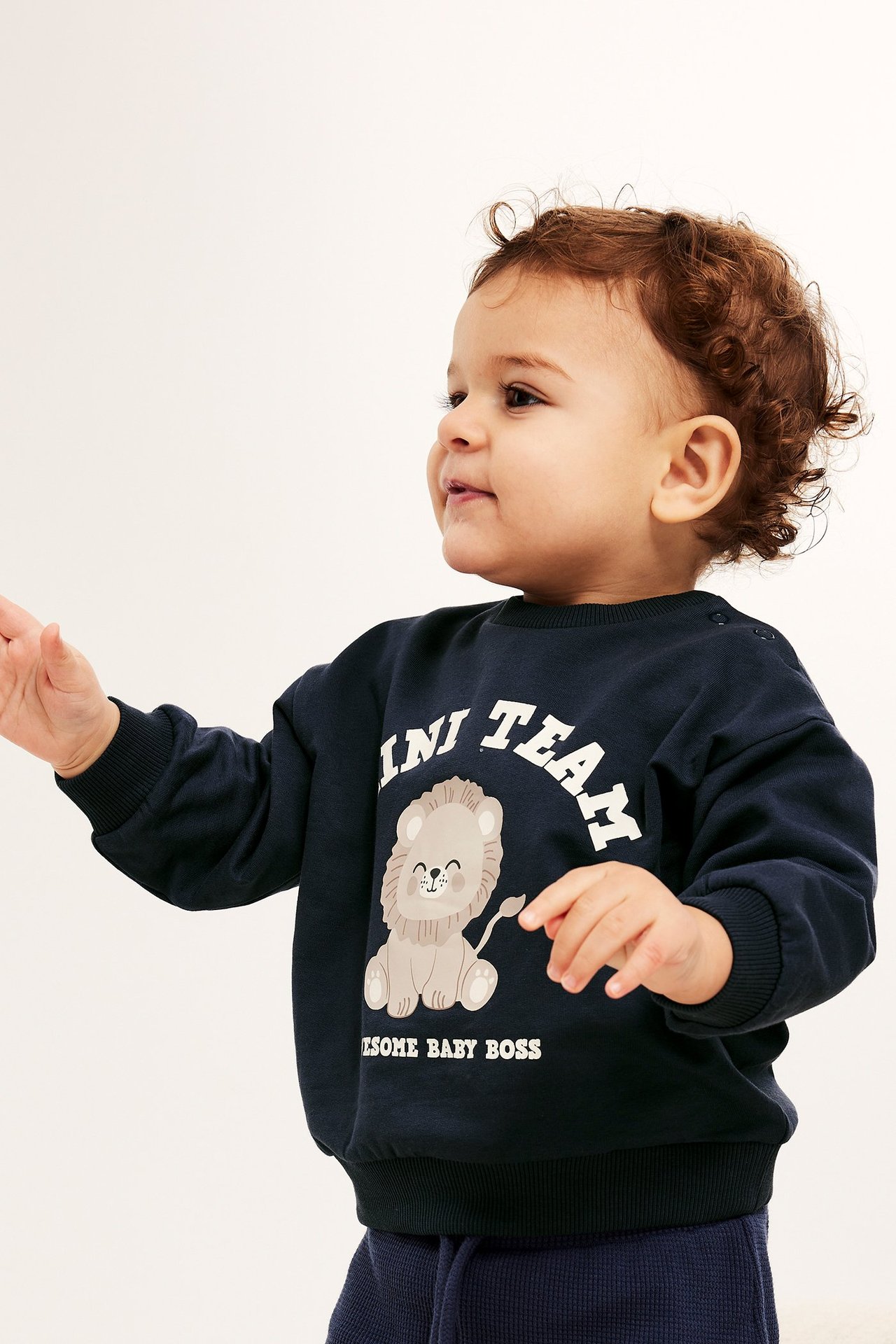 Sidevisning av baby i mørkeblå genser med løvetrykk og skulderknapper, kombinert med ribbestrikket bukse.