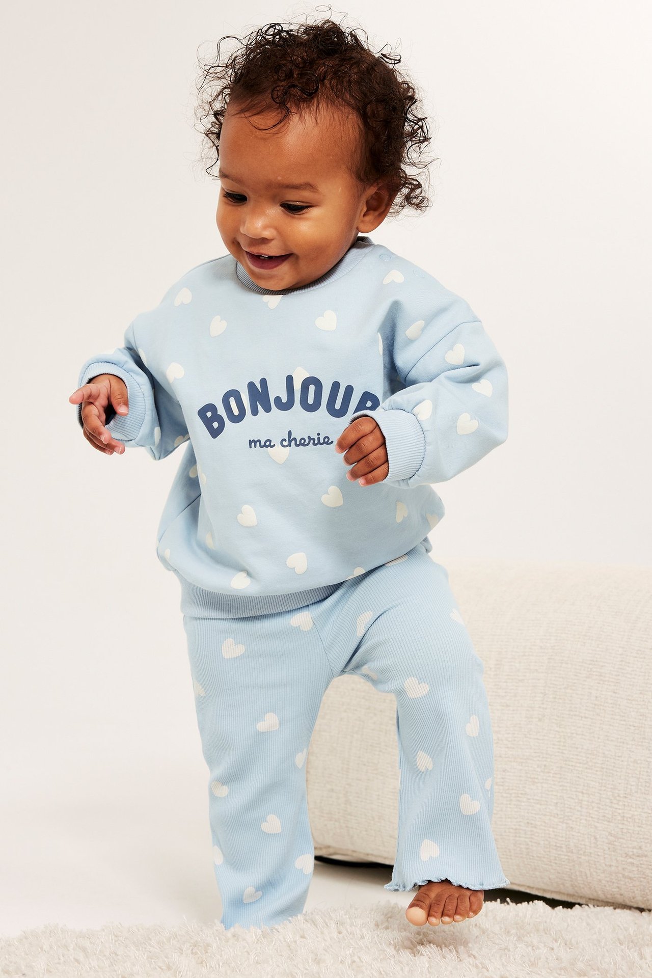 Frontbilde av baby i lyseblå mønstret sweatshirt med tekst og matchende ribbestrikket bukse.