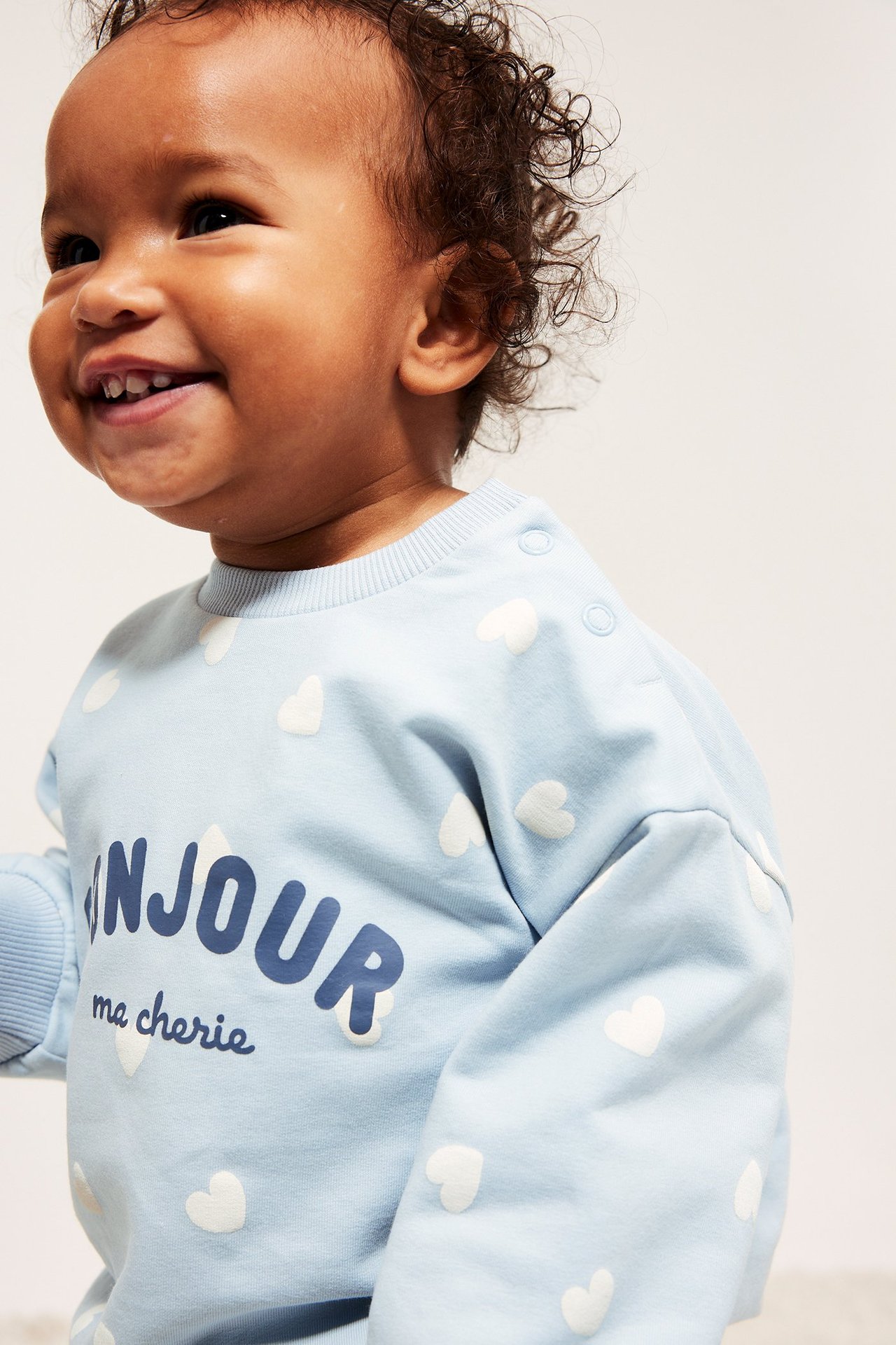 Nærbilde av en baby i lys blå sweatshirt med hvitt hjerteprint, tekst og skulderknapper.