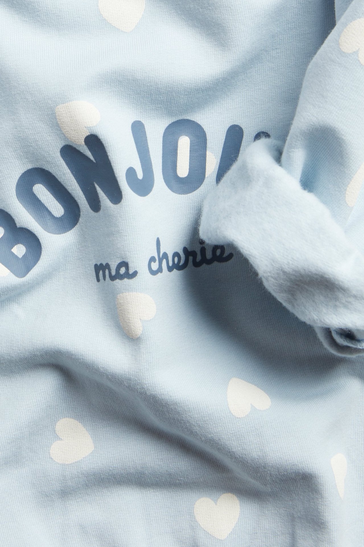 Zbliżenie na jasnoniebieską bluzę niemowlęcą z białymi sercami i napisem 'BONJOUR ma cherie'.