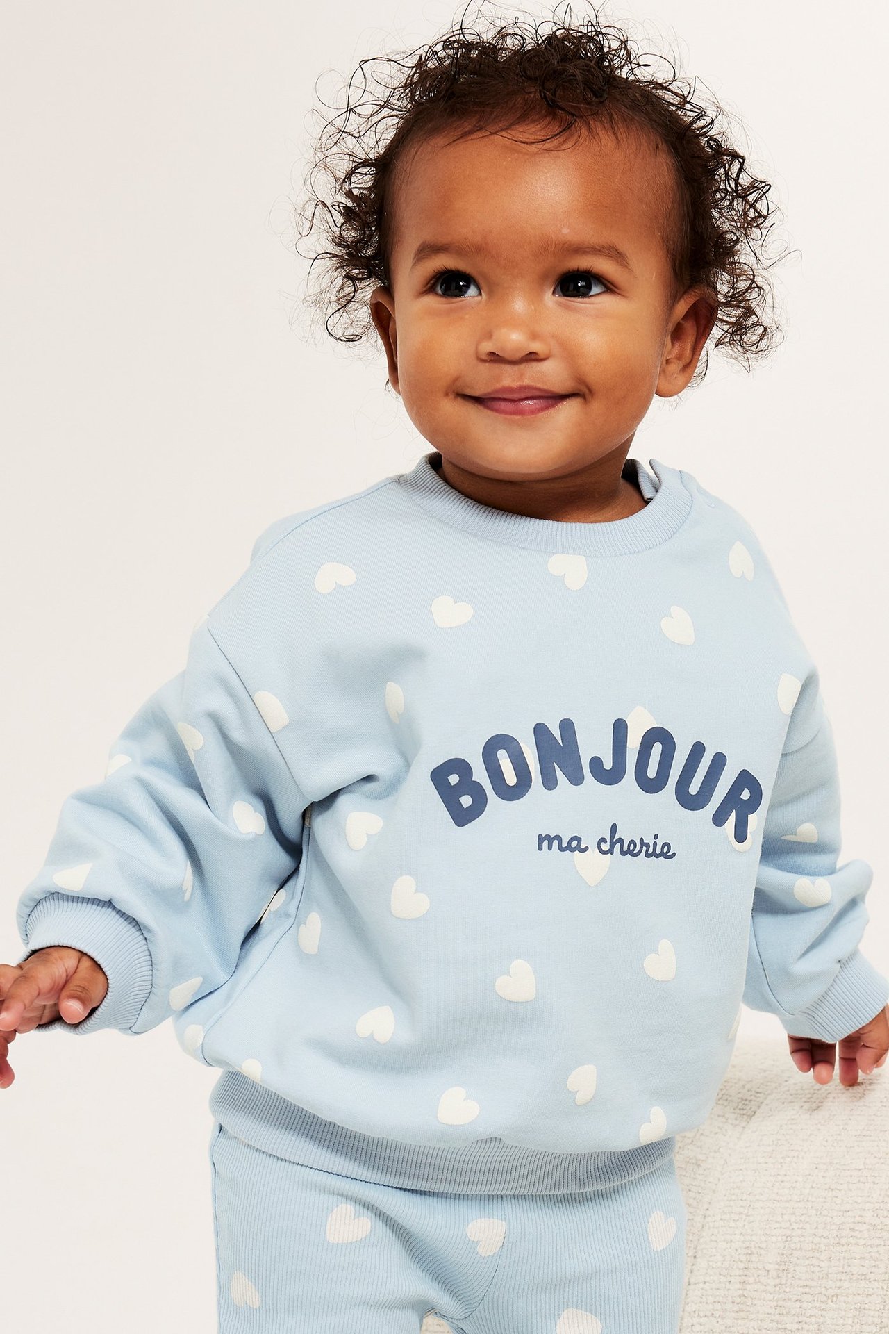 Front: baby i lyseblå sweatshirt med hvite hjerter, "Bonjour ma cherie" trykk og matchende bukse.