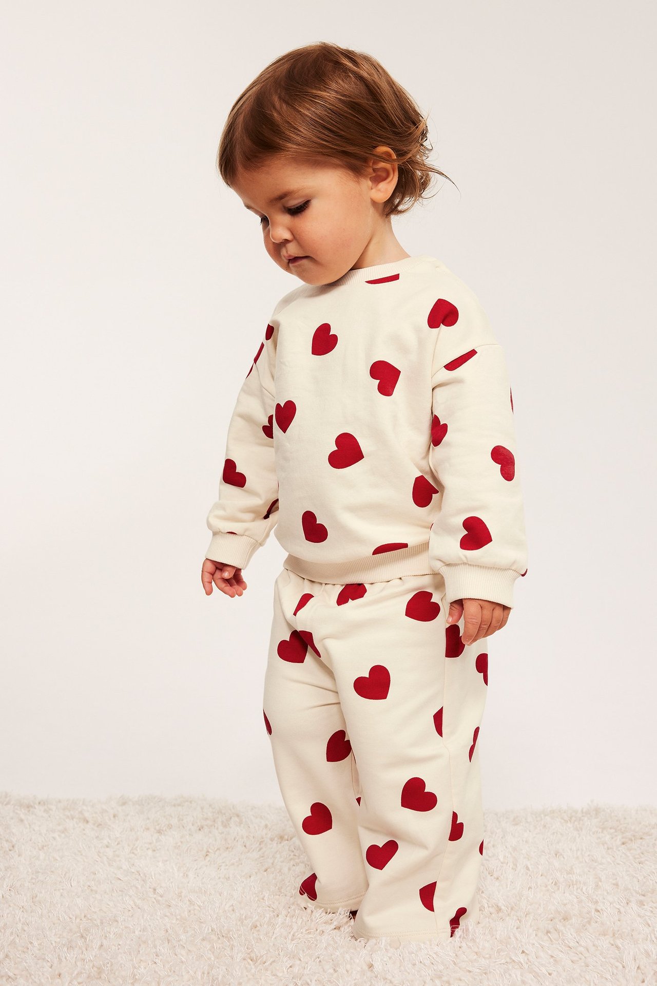 Sidebilde av baby i offwhite bomullssweatshirt med rødt hjerteprint og matchende bukser.