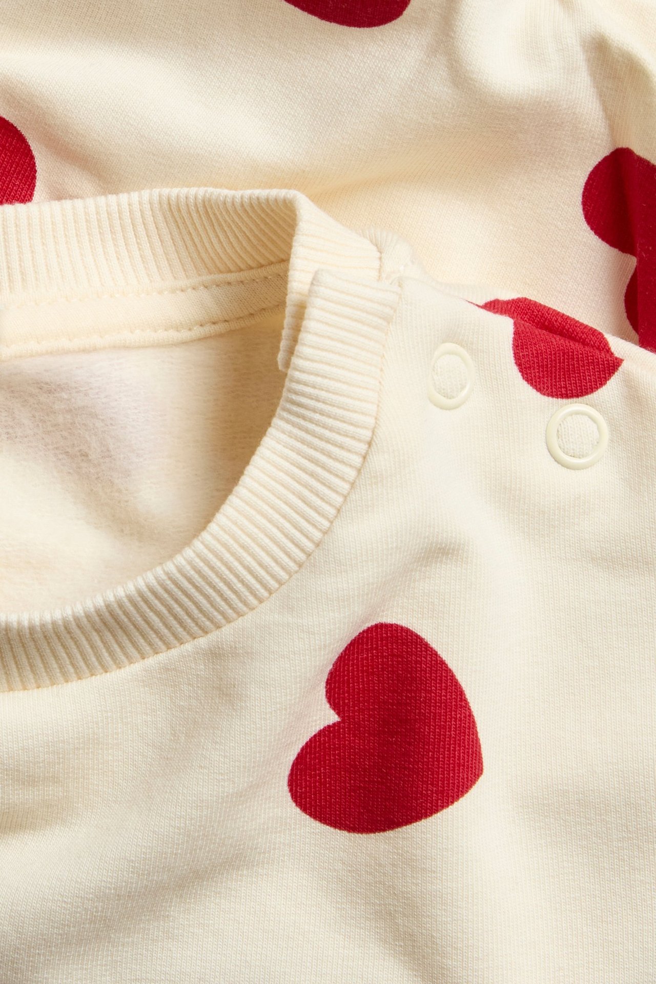 Nærbilde av en offwhite babygenser med rødt hjerteprint, ribbet hals og trykkknapper på skulderen.