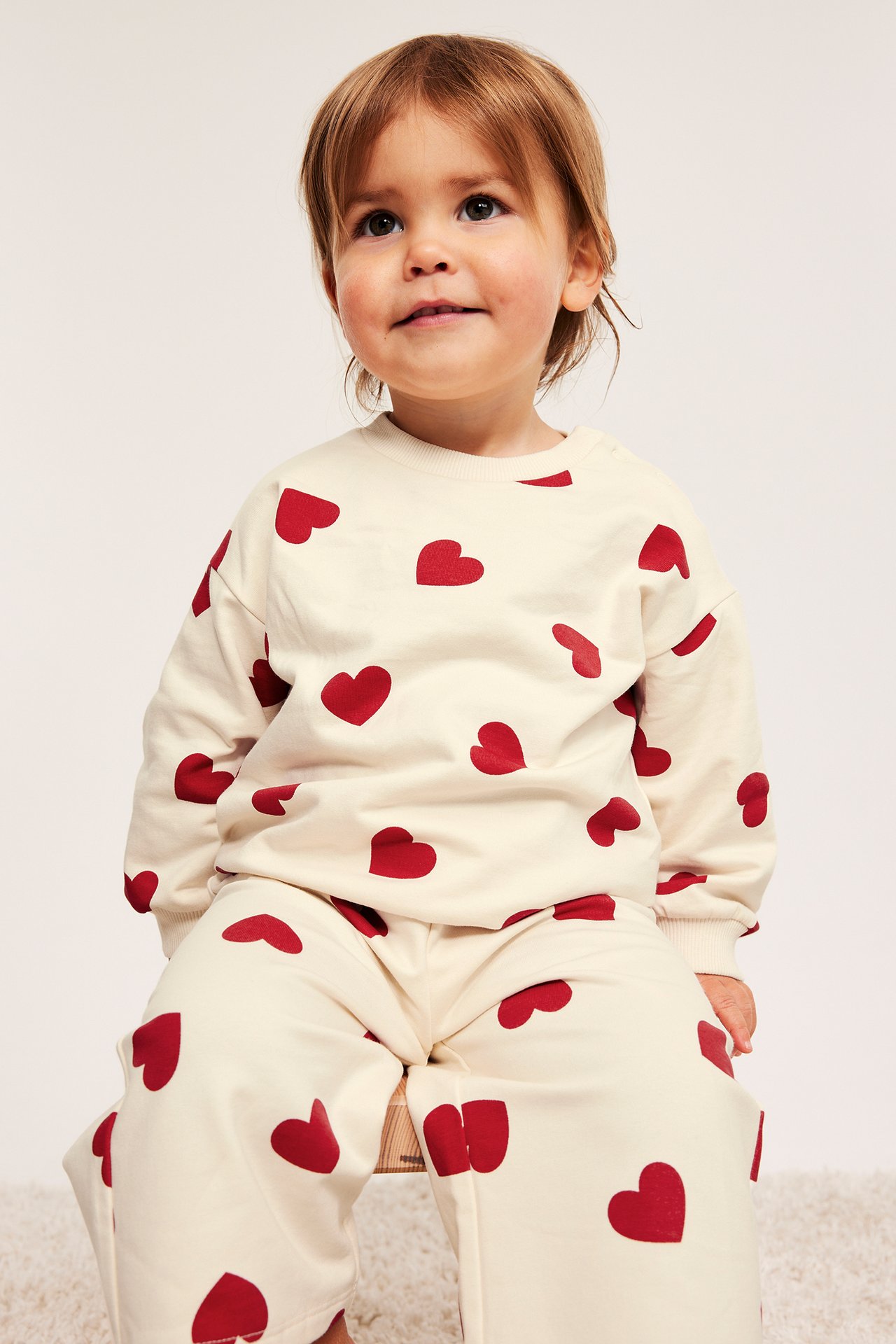 Baby har på offwhite sweatshirt med røde hjerter og matchende joggebukser, frontvisning.
