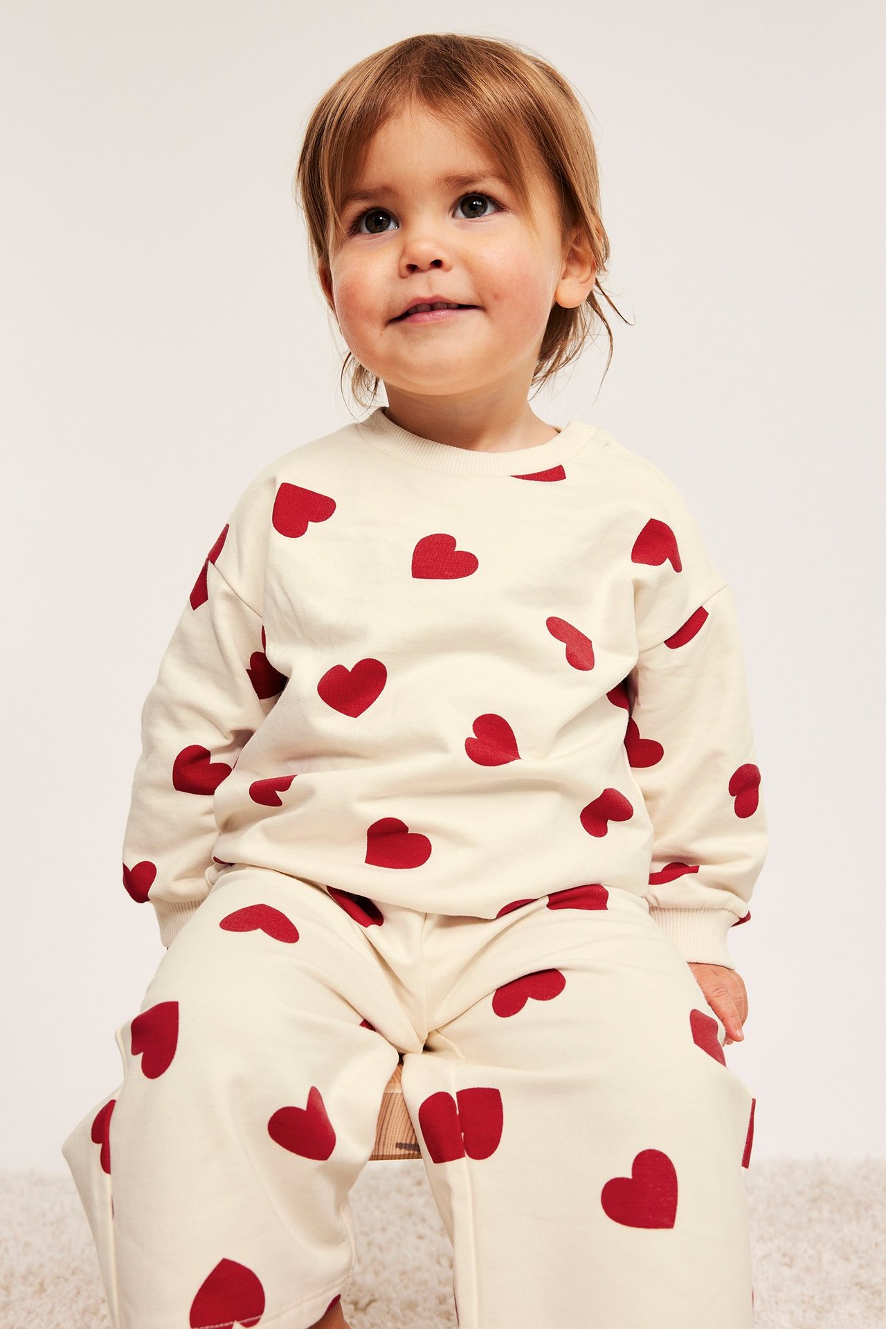 Baby har på offwhite sweatshirt med røde hjerter og matchende joggebukser, frontvisning.