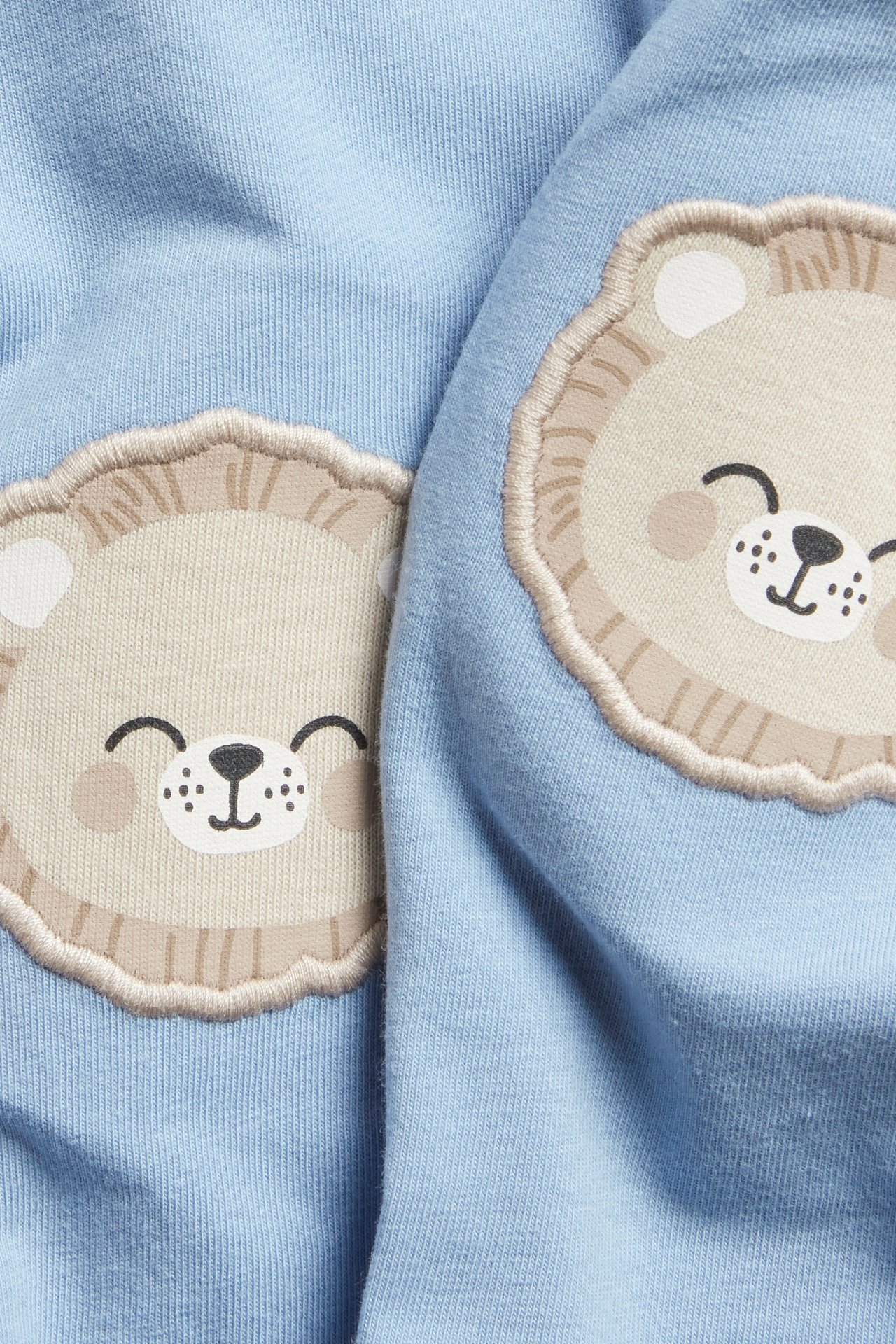 Nærbilde av lyseblå baby leggings med to beige løveansikt-applikasjoner på knærne.