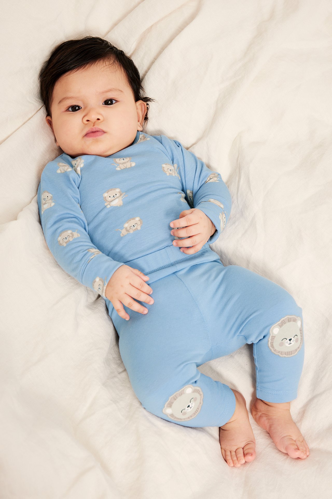 Z przodu: Niemowlę w jasnoniebieskich legginsach z aplikacjami lwów na kolanach i pasującej bluzce w lwy.