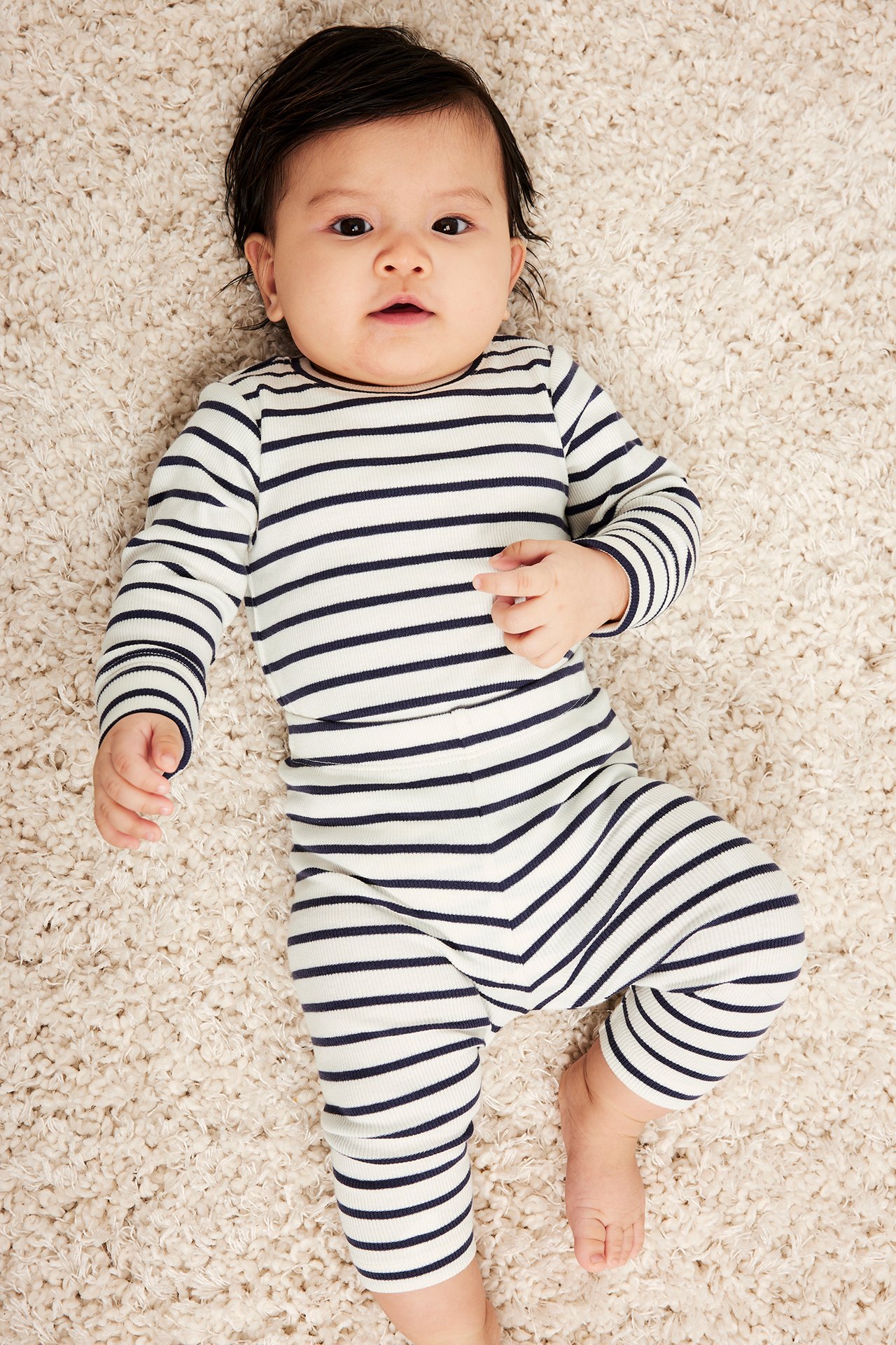 Framifrån, baby i långärmad, långbent, ribbad vit body med mörkblå horisontella ränder.