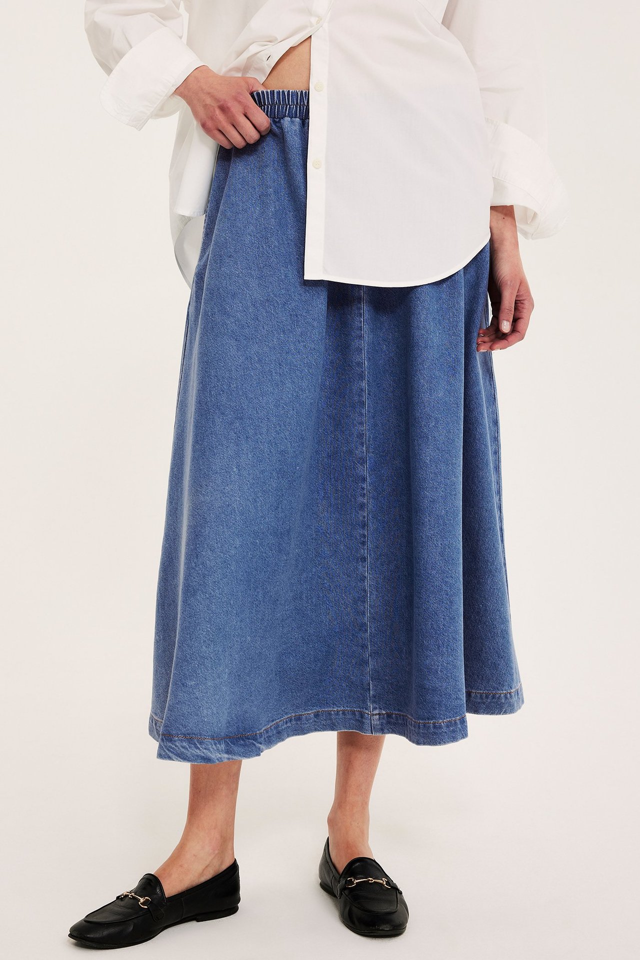 Framifrån: Kvinna i blå denim A-linjeformad midikjol med resår, vit skjorta och svarta loafers.