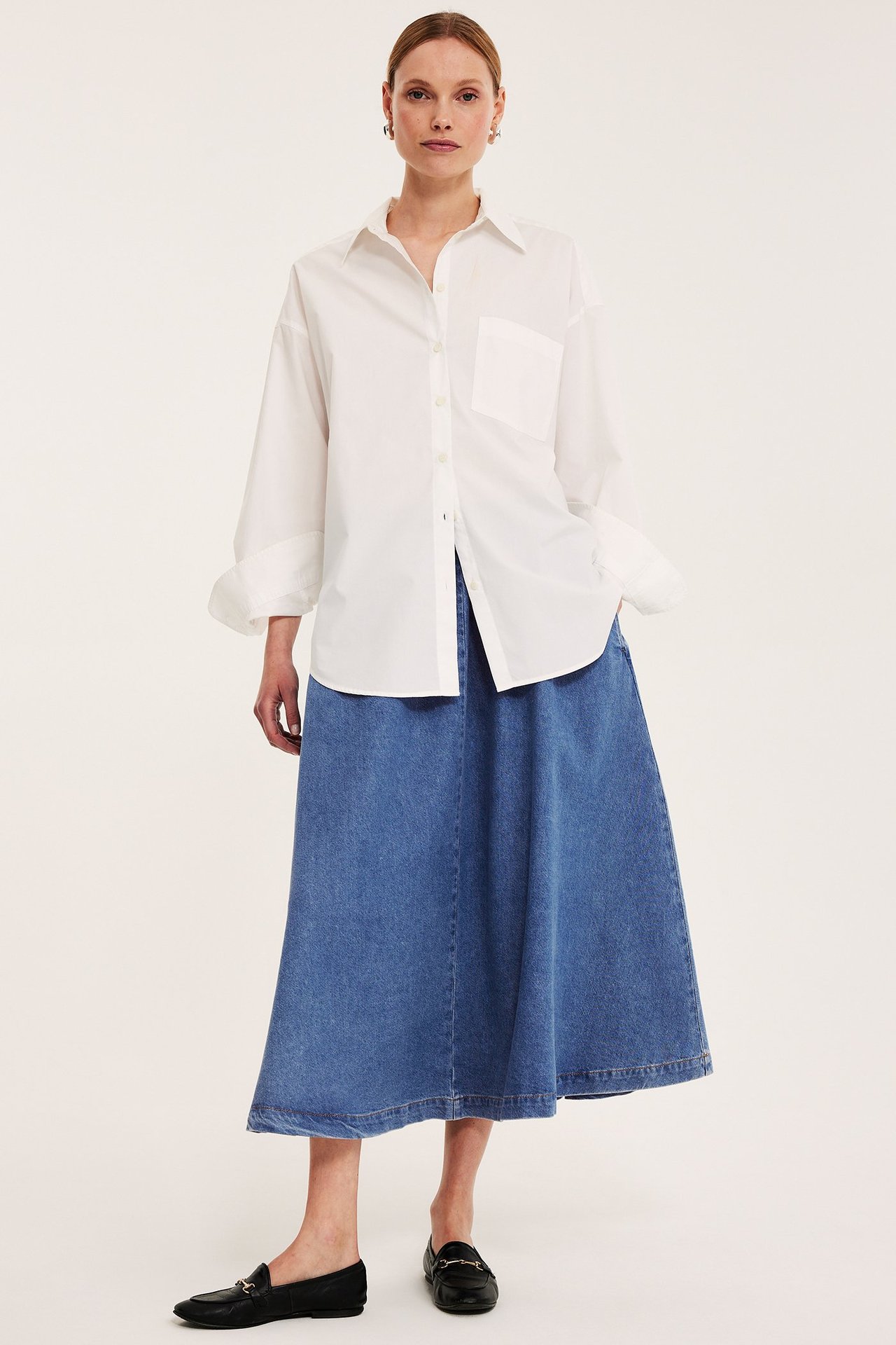 Framifrån: kvinna i lång blå A-linjeformad jeanskjol, vit oversized skjorta och svarta loafers.