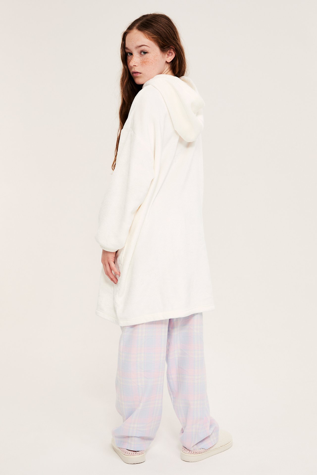 Bakfra: Et barn i en oversized offwhite fleecehoodie, rutete vide bukser og lyse tøfler.