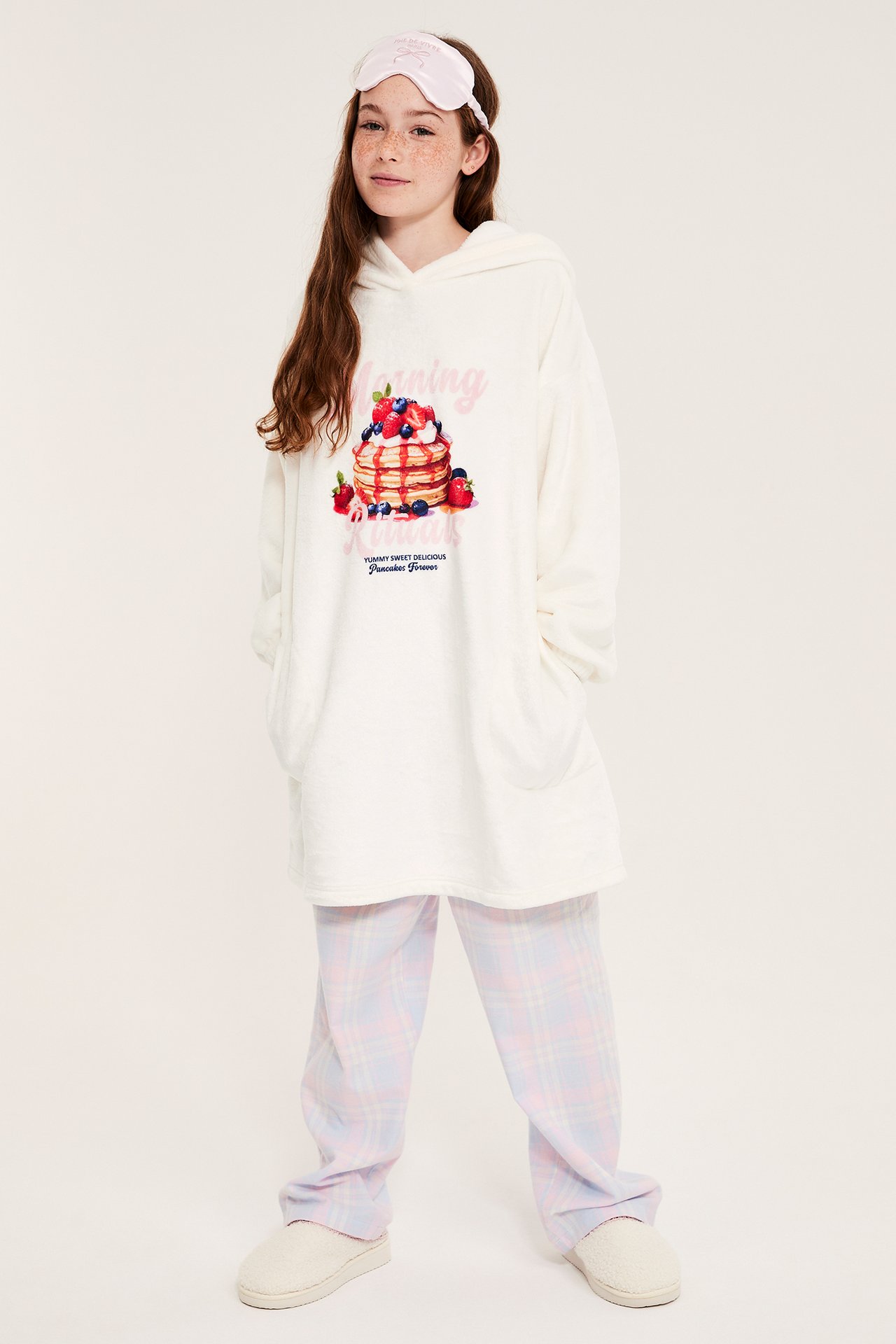 Front: Barn i offwhite fleecegenser med hette og pannekaketrykk, rutete pyjamas, beige tøfler, rosa sovemaske.