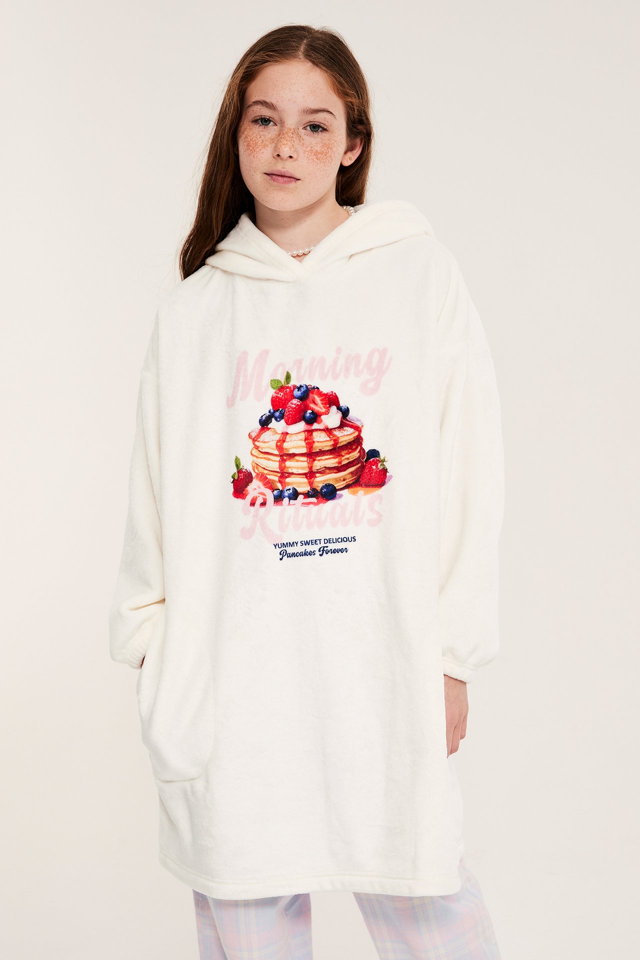 Framifrån: Barn i en naturvit oversize fleecehoodie med pannkaksprint, händerna i fickor, över rutiga byxor.
